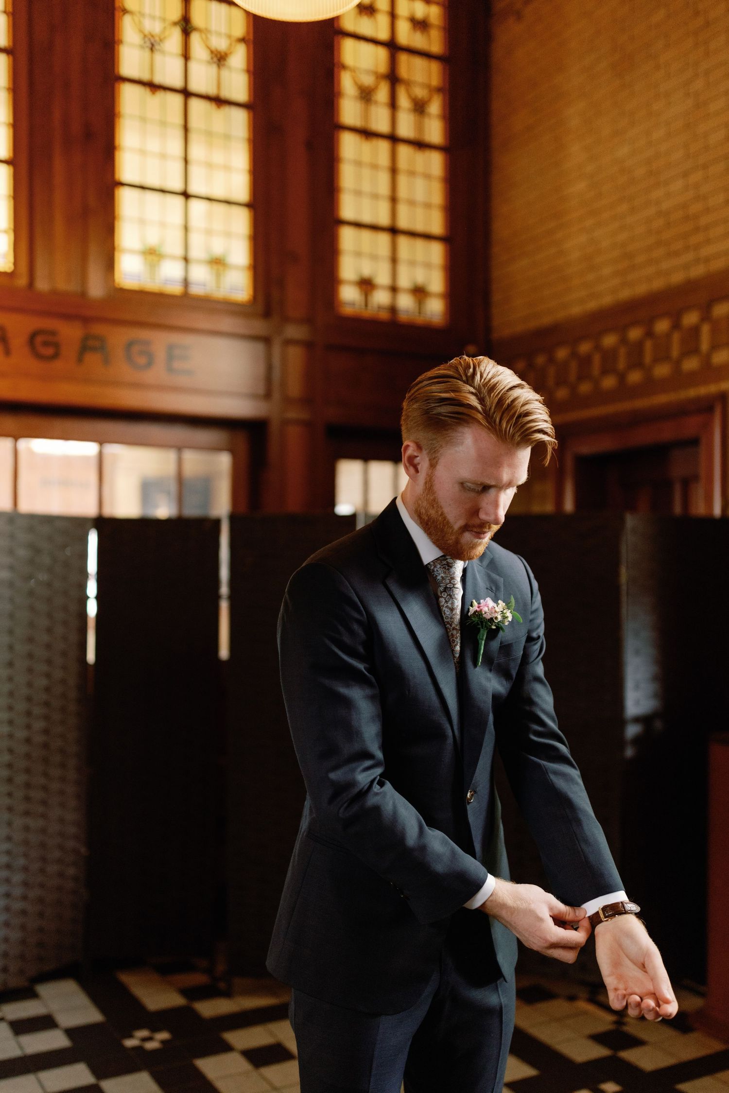Man in black suit checks his watch while standing in a dimly lit room with industrial windows.