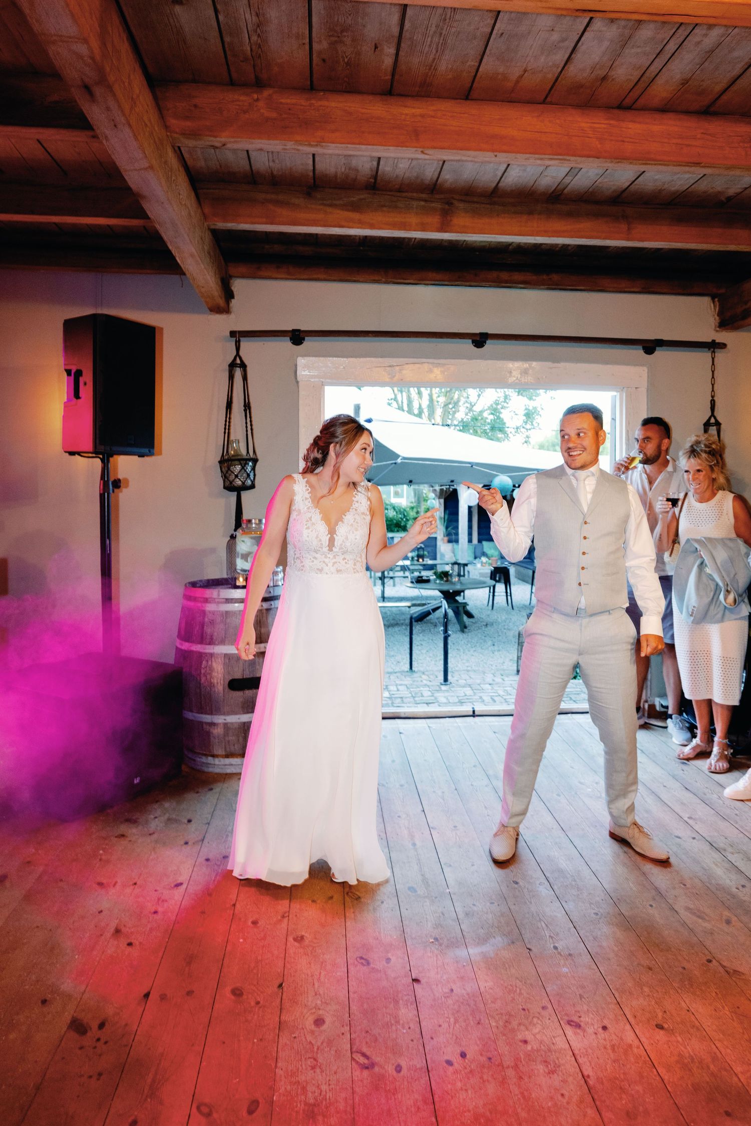 Couple dancing on rustic wooden floor under exposed beam ceiling with pink and purple smoke effects during wedding celebration.