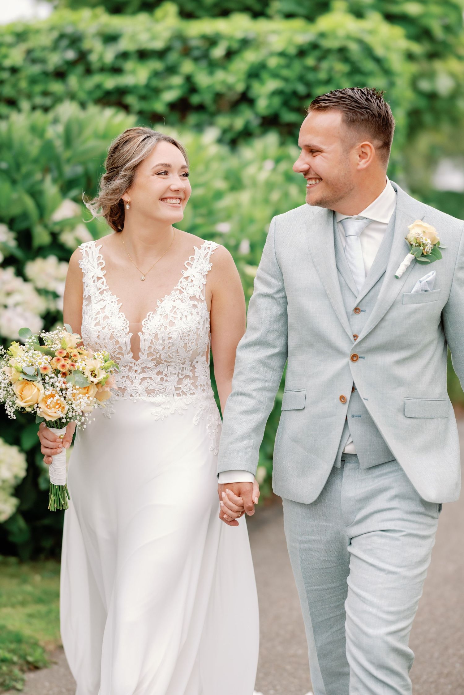 A couple in wedding attire walking and smiling together in a garden setting with peach-colored bouquet details.