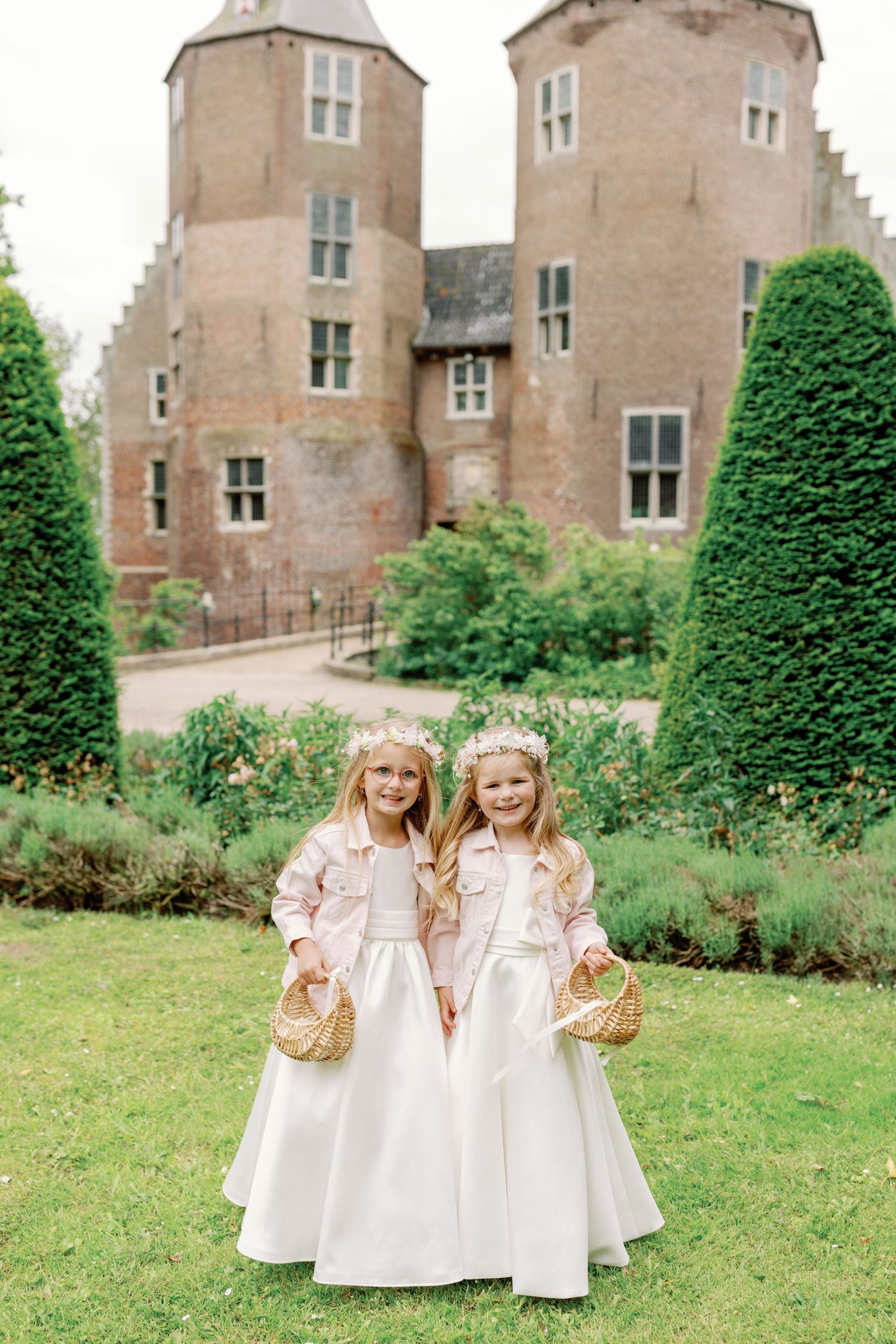 Two flower girls in white dresses play on the castle grounds with manicured topiaries and stone walls in the background.