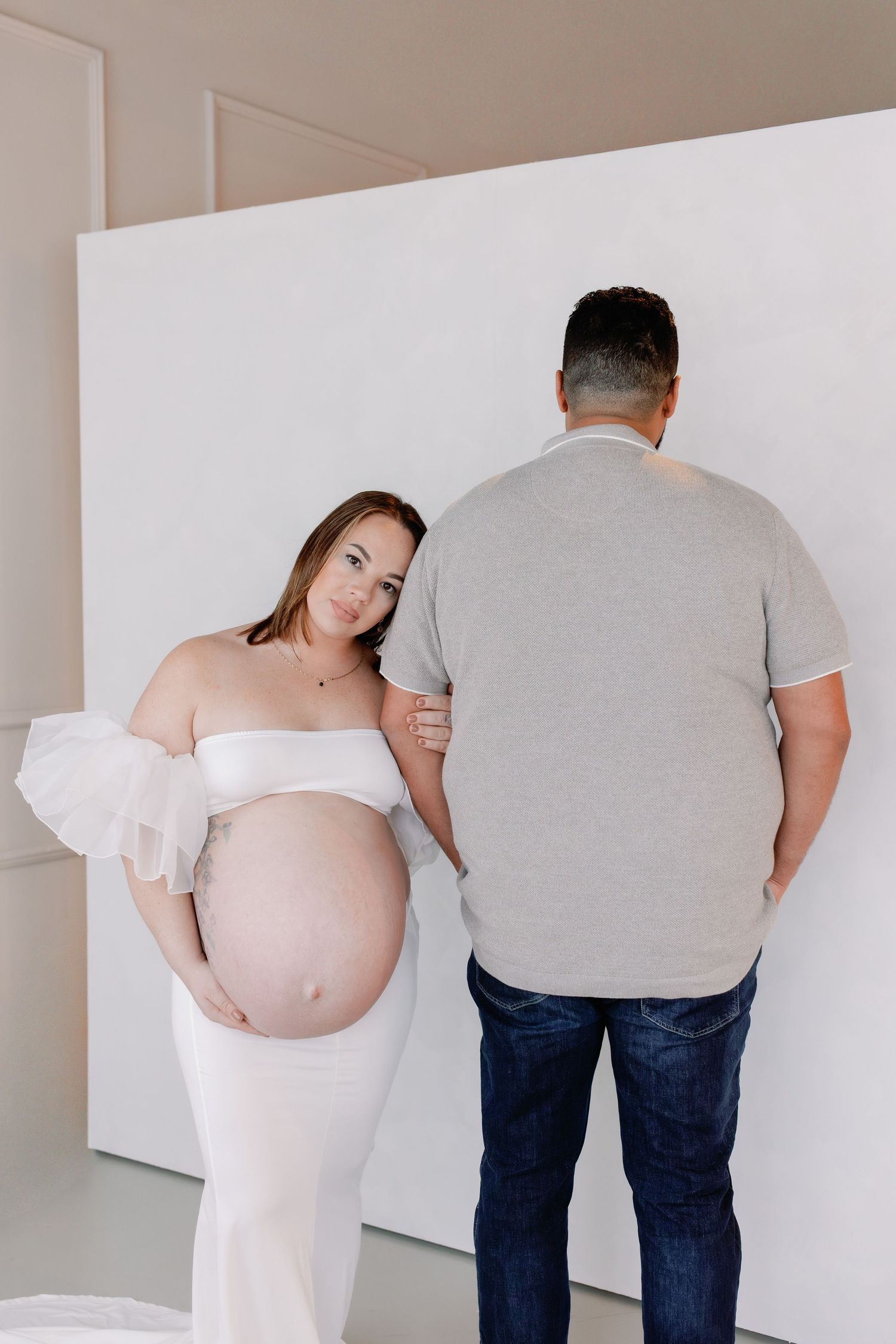 A creative pregnancy photo series showing multiple poses of an expecting couple in white and casual attire against a white wall.