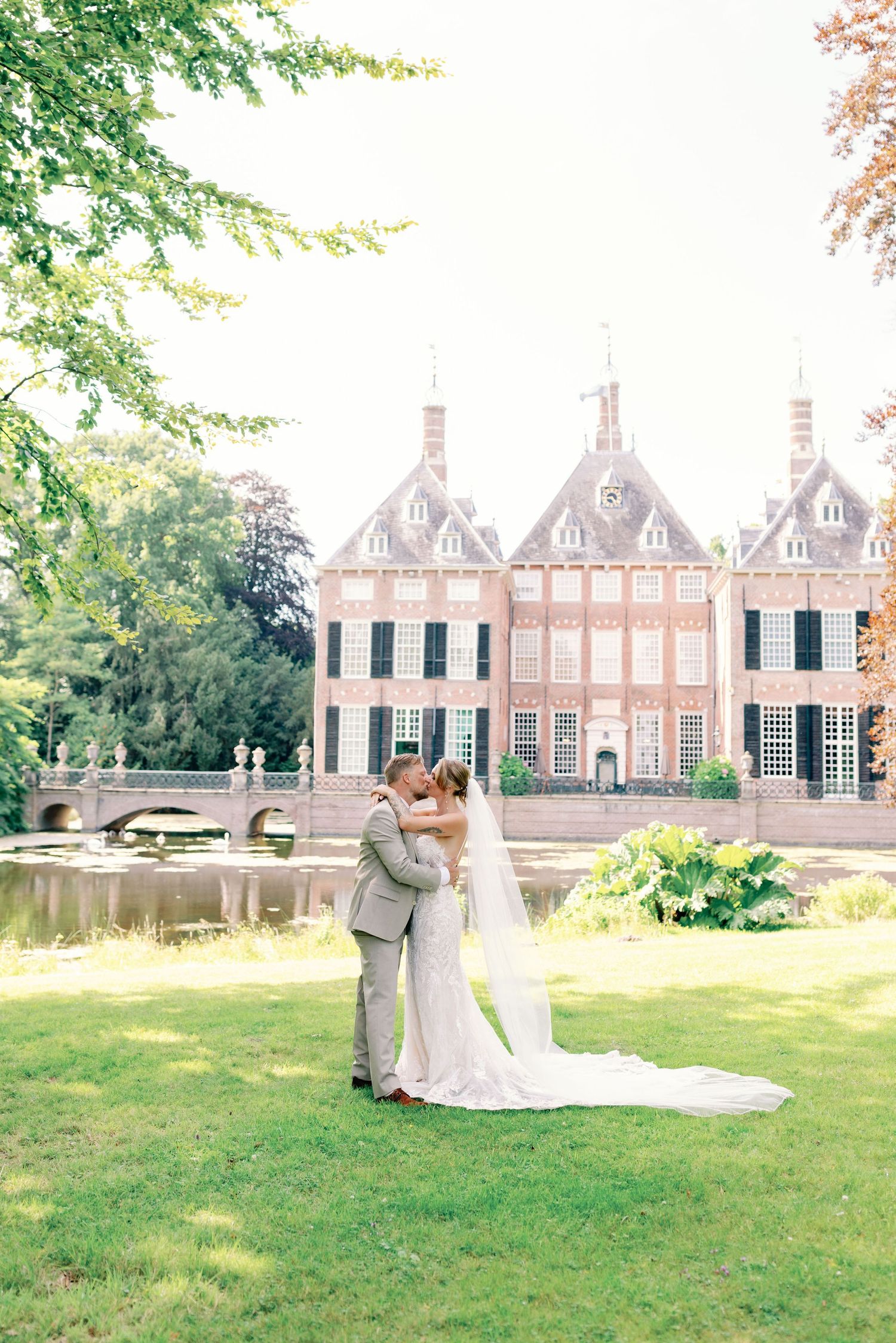 A wedding couple poses in front of a grand historic brick mansion with manicured gardens.