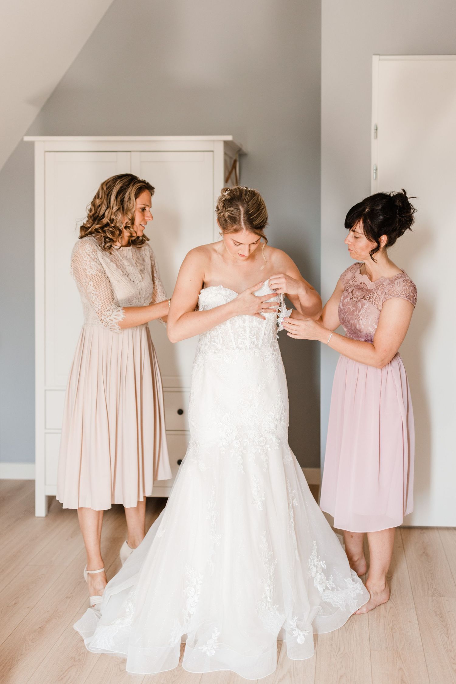 Wedding party helps bride with final dress adjustments in a light-filled room with pale pink bridesmaid dresses.