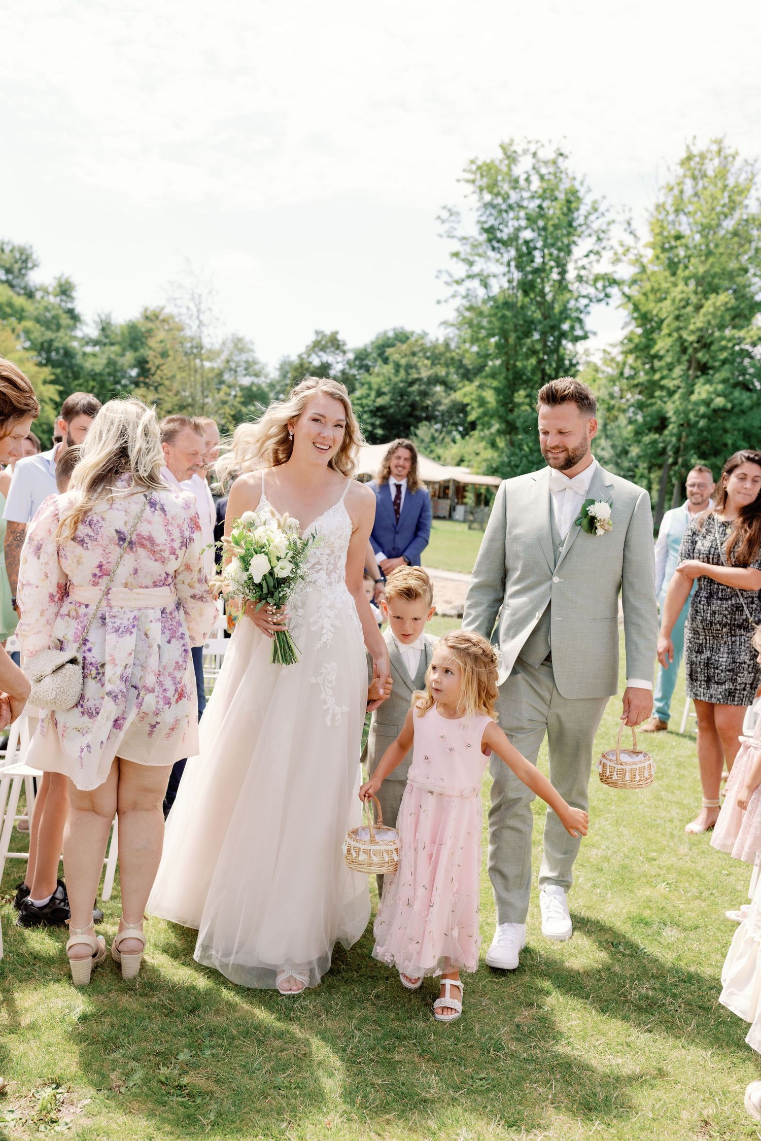 Wedding party celebrates outdoors on sunny day with guests in floral dresses and light summer attire.