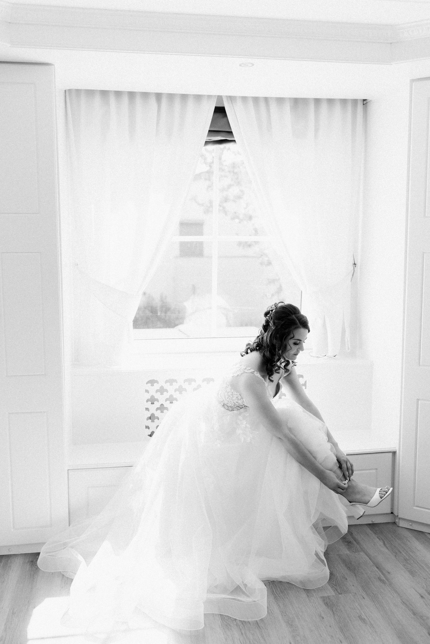 Black and white photo of someone in a flowing white wedding gown adjusting their shoe near a bright window.