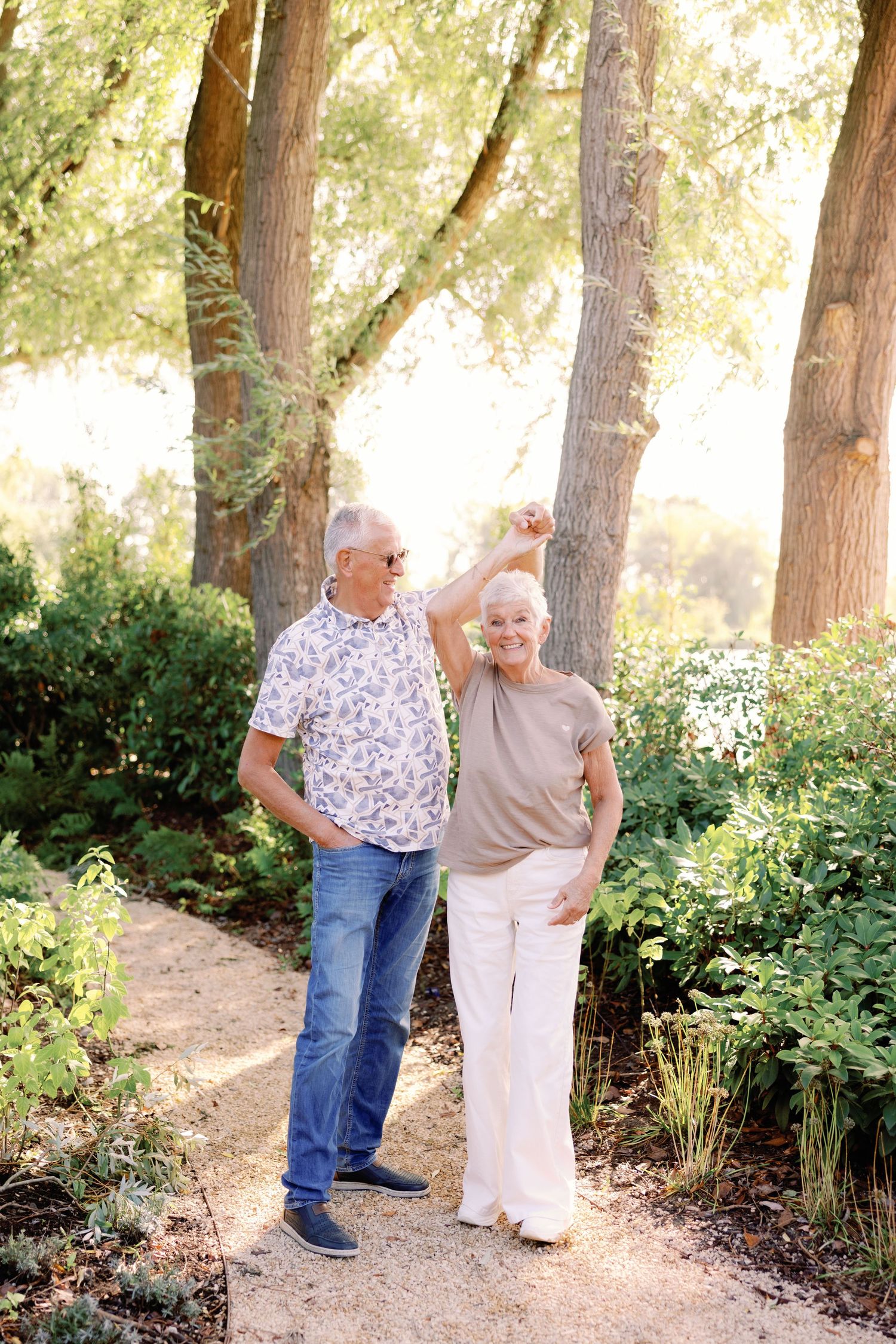 Smiling older couple dancing together on a sunlit garden path surrounded by trees and lush greenery.