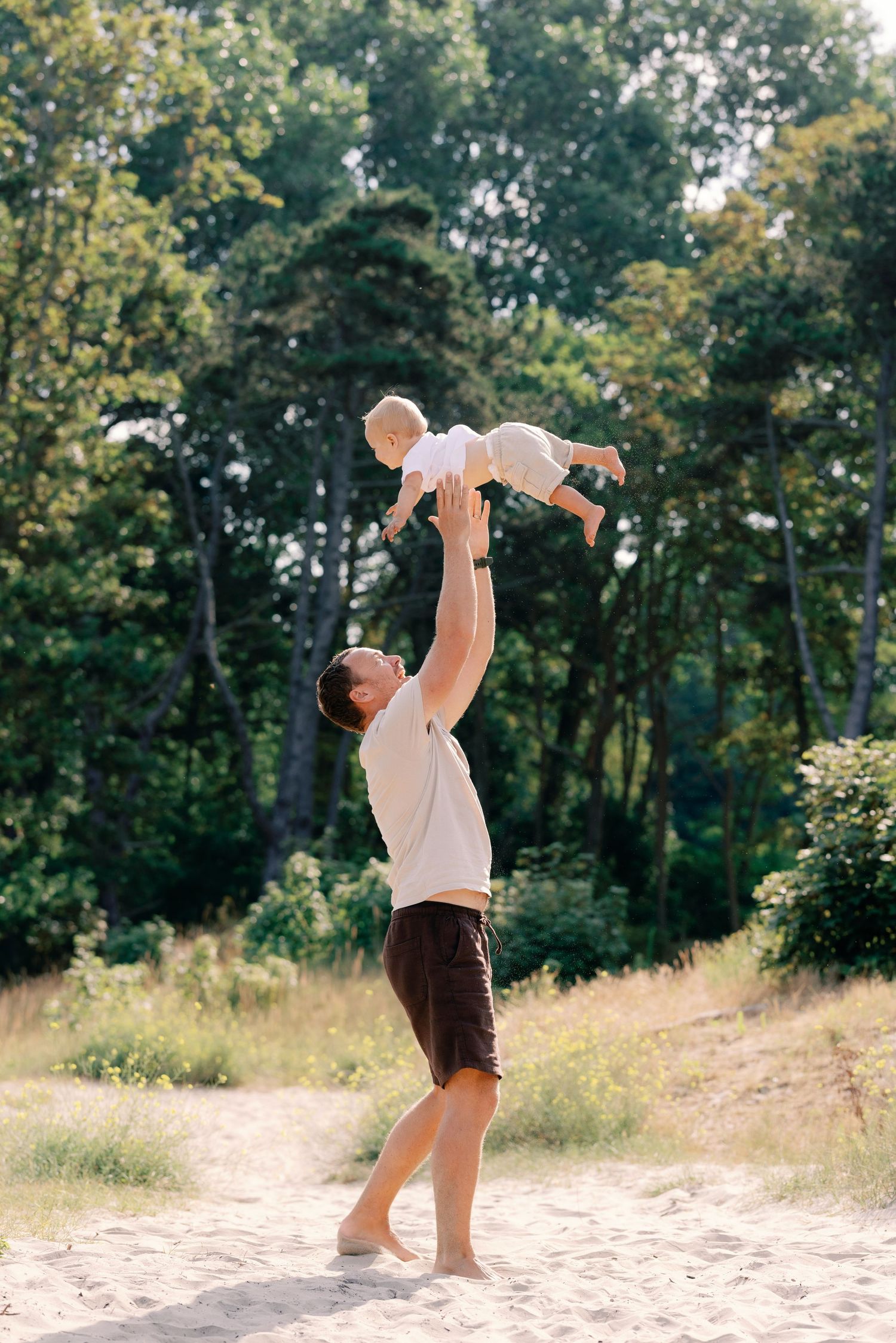 Father joyfully lifting baby high in the air outdoors on sandy ground with lush green trees in background.