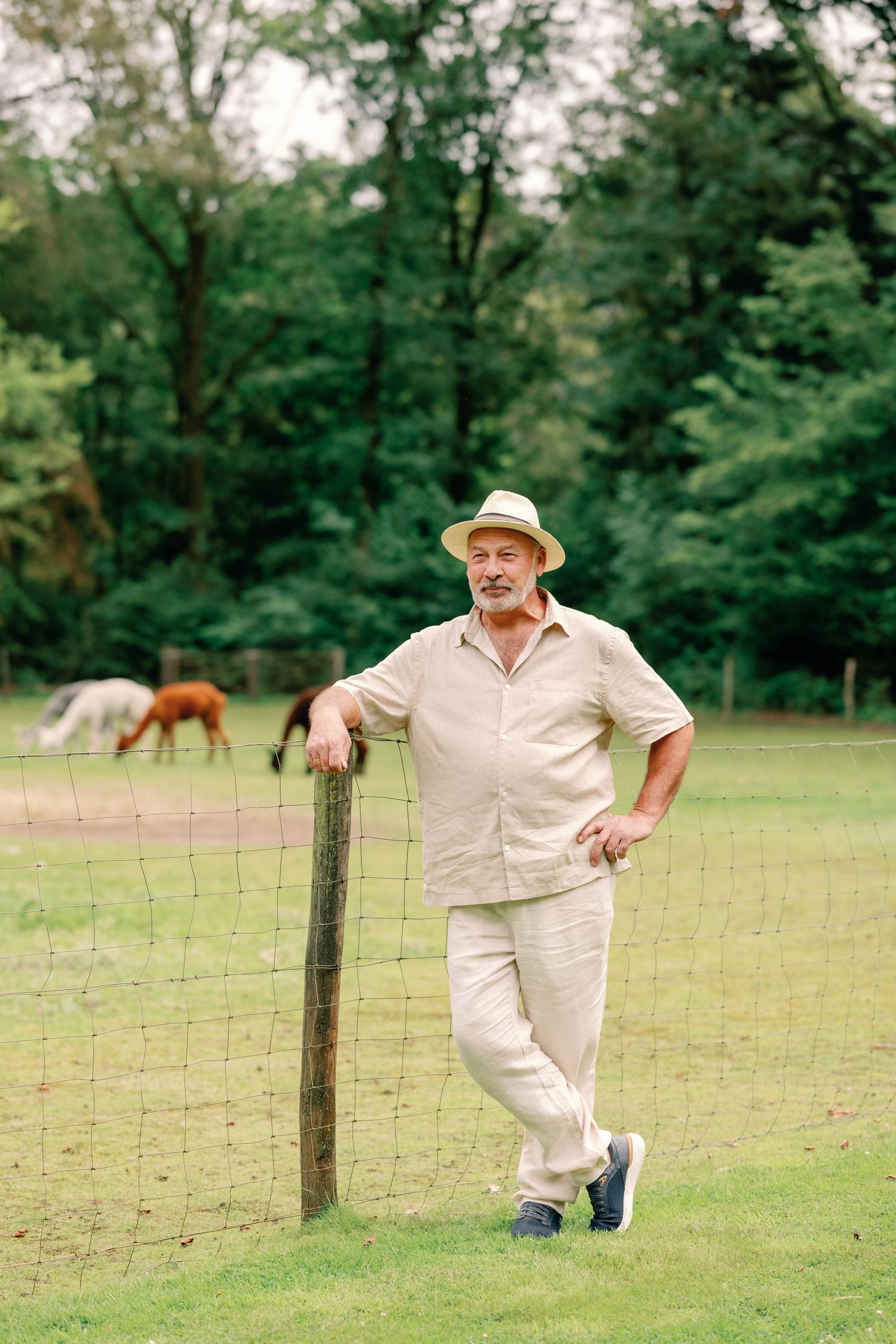Person in light clothing and hat leaning against a farm fence with horses in background.