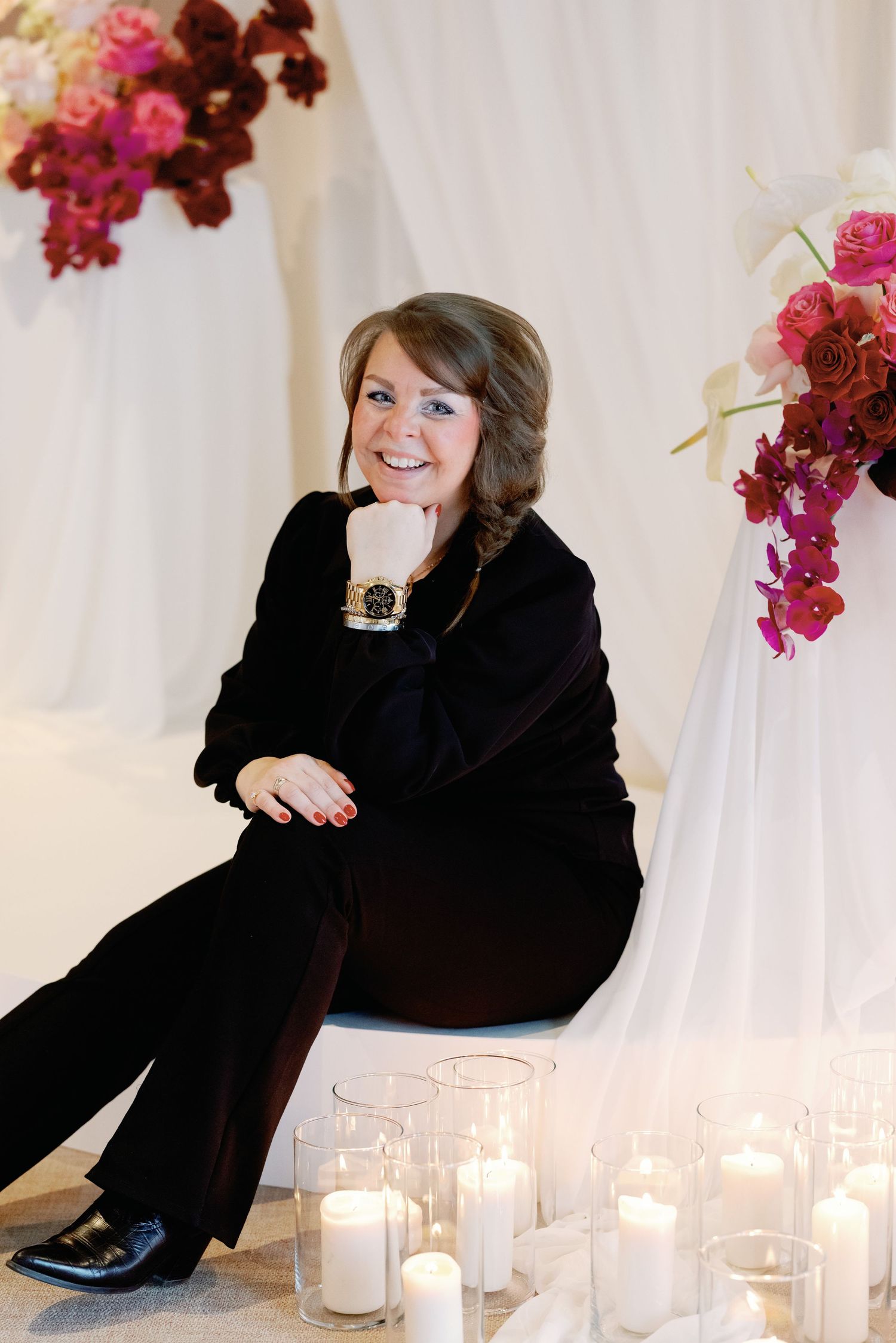 A person in black attire sits surrounded by white candles and pink floral decorations in an elegant setting.
