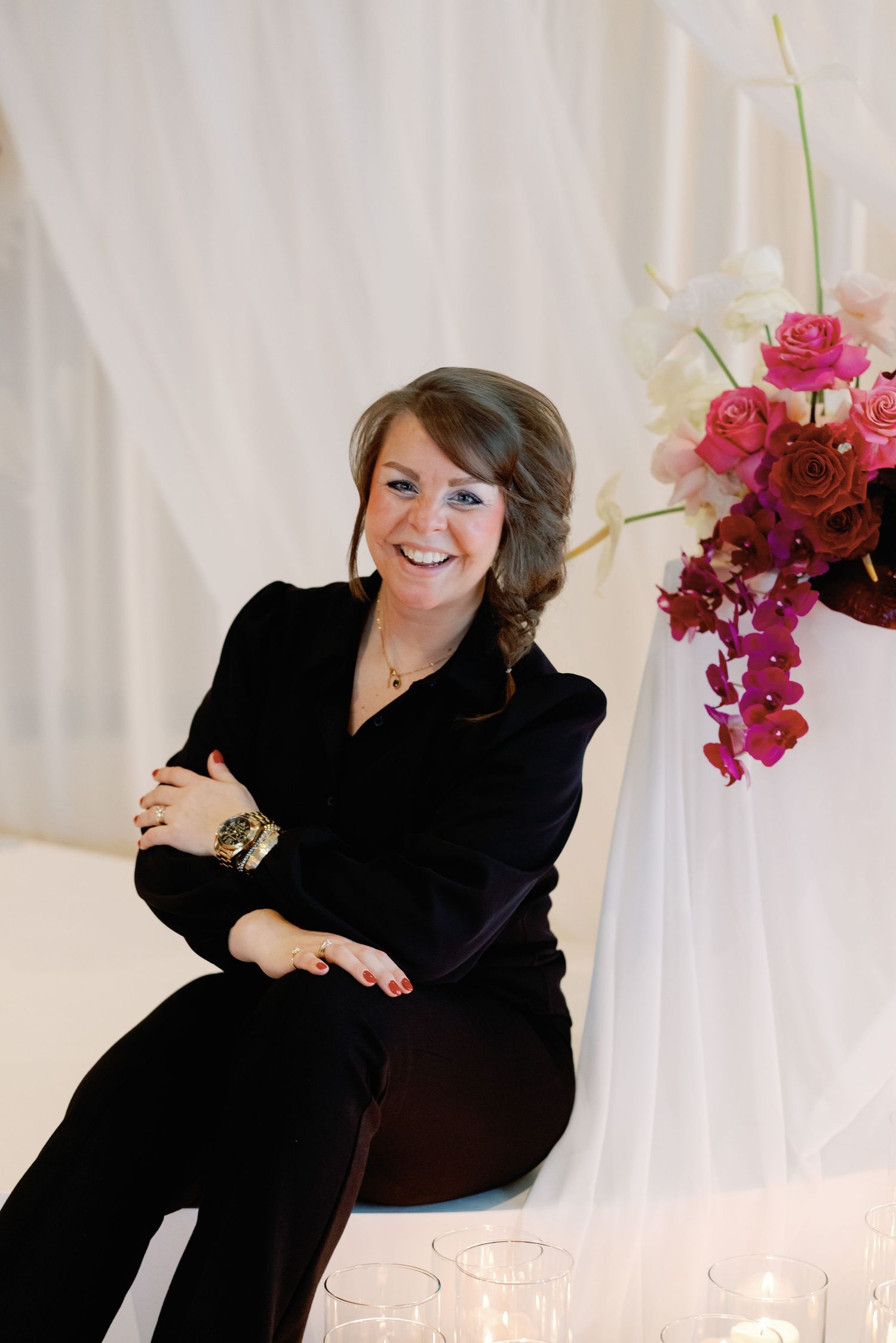 Professional portrait with elegant black attire seated beside floral arrangement of roses and orchids against white backdrop.