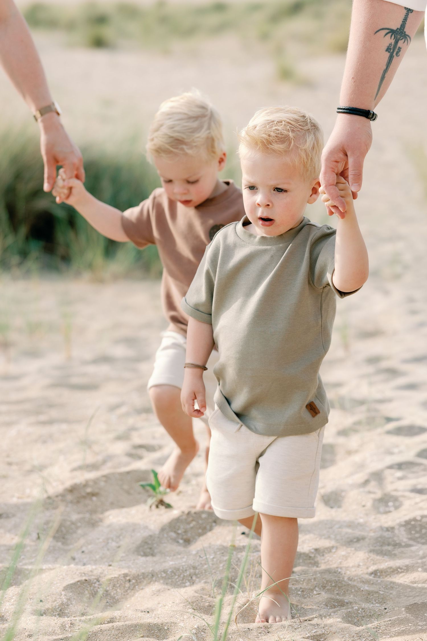Two toddler boys in matching earth-tone outfits walk hand-in-hand with adults on a sandy beach.