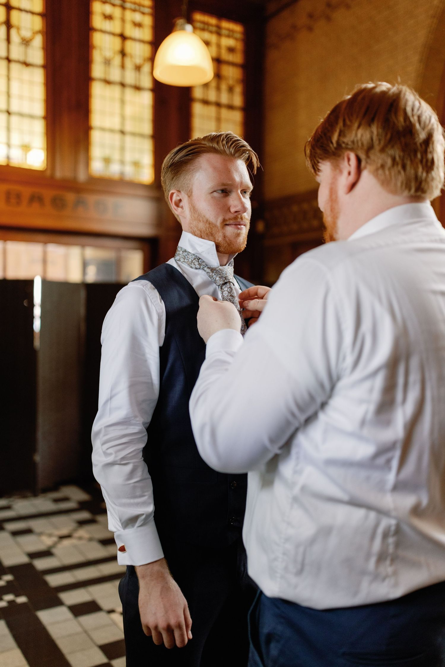 Two people in formal attire share a candid moment in a warmly lit room with vintage windows.