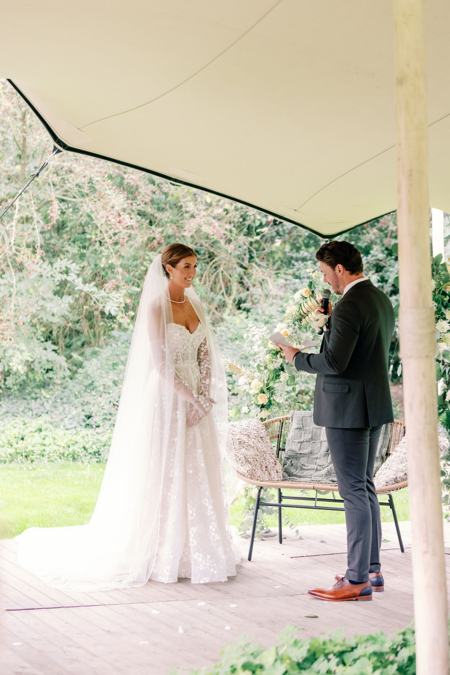 A couple exchanges vows during their outdoor wedding ceremony under a white gazebo surrounded by lush greenery.