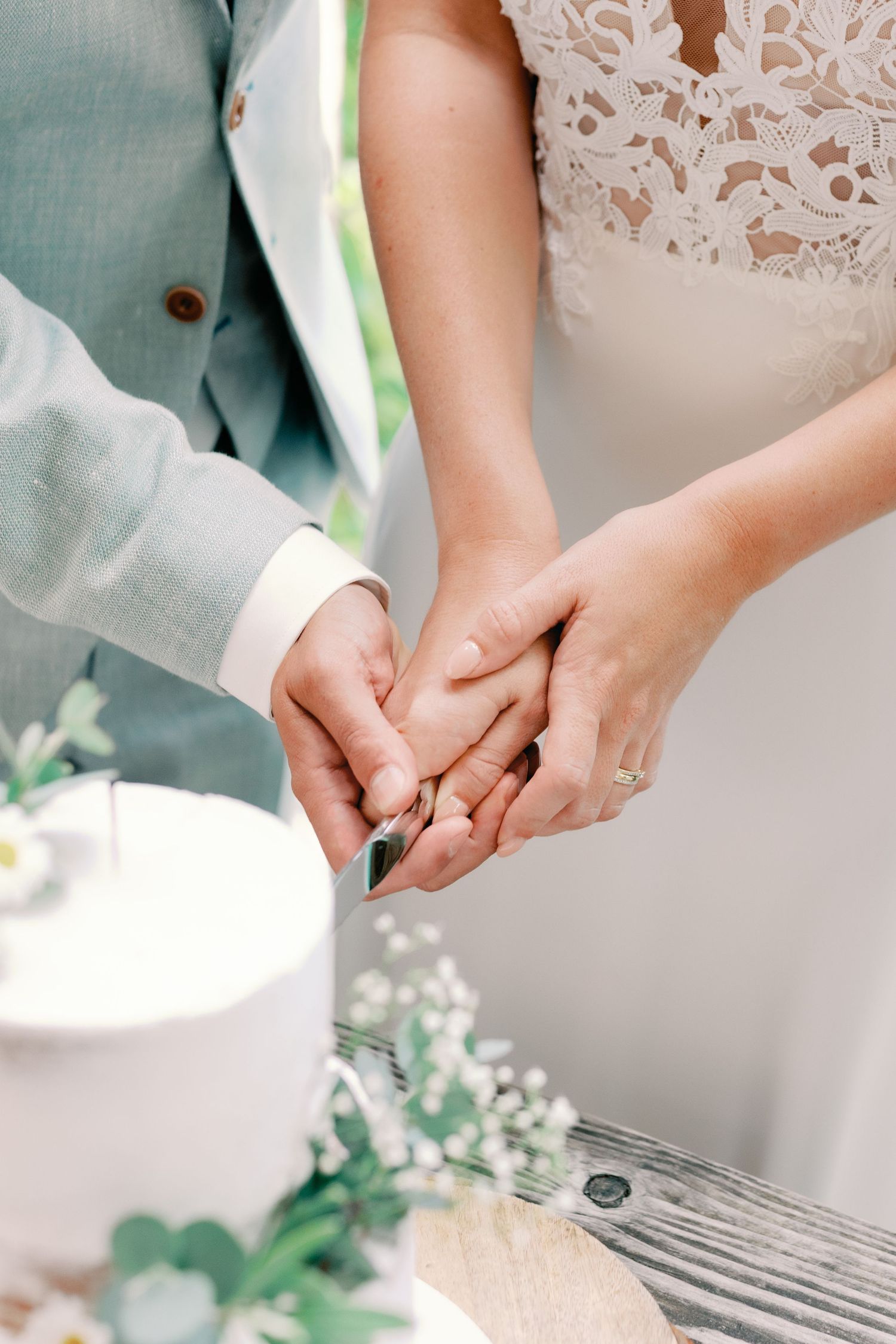 Close-up of newlywed couple holding hands, showing wedding rings against white lace dress details.