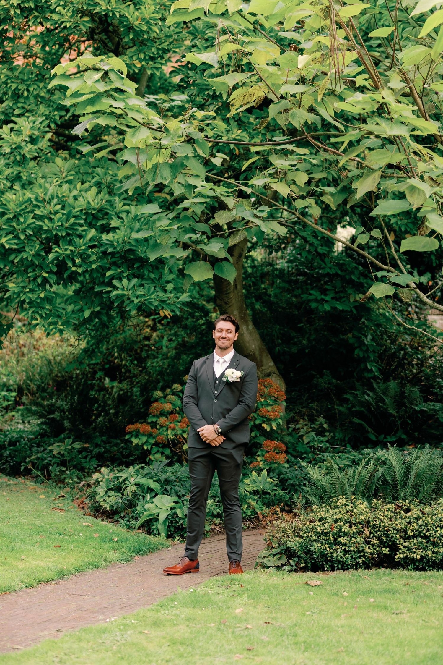 A person in a grey suit and boutonniere stands on a garden path surrounded by lush greenery and flowering shrubs.