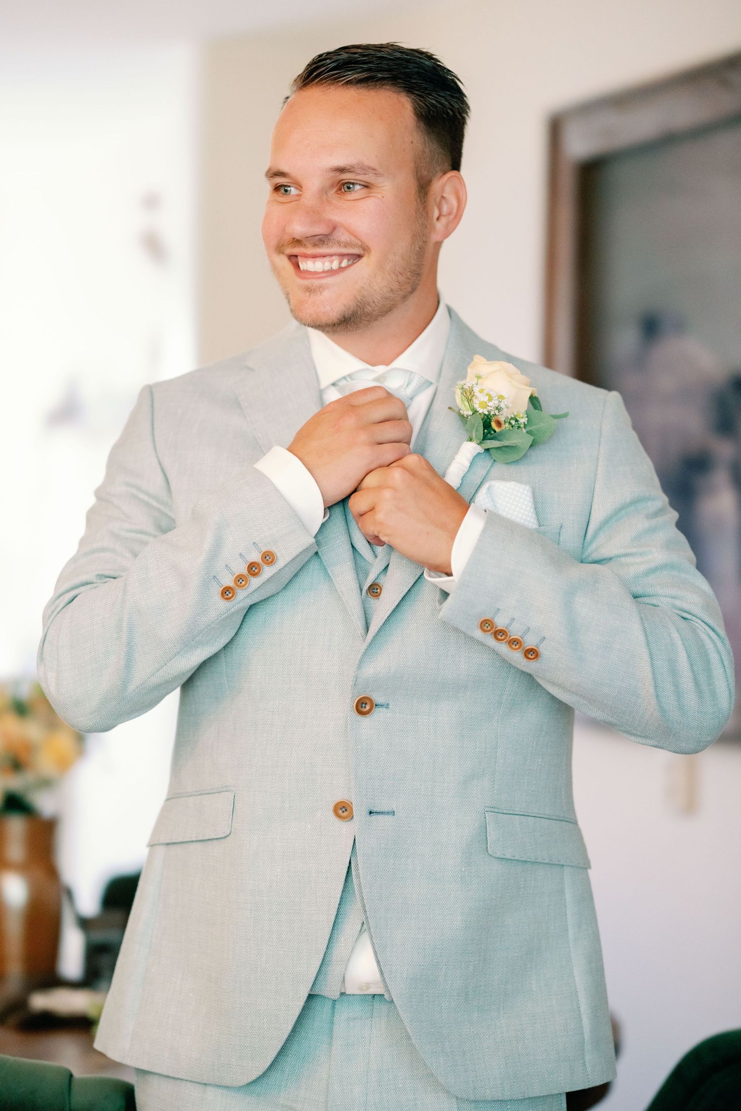 A groom in a light gray suit adjusts his tie while smiling during wedding preparations.