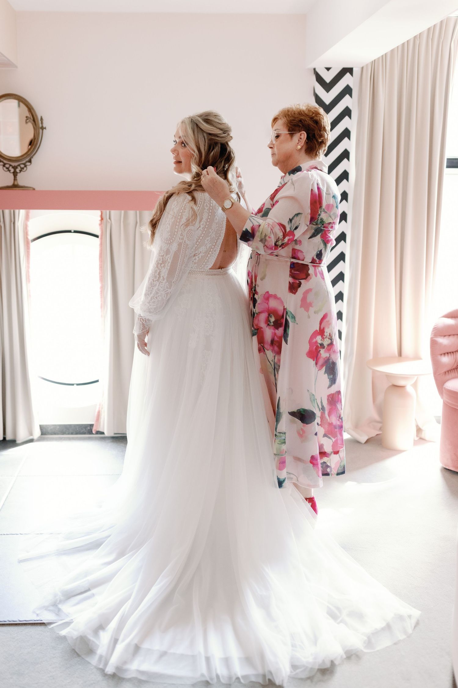 A bride in a flowing white wedding gown stands beside someone wearing a pink floral dress during wedding preparations.