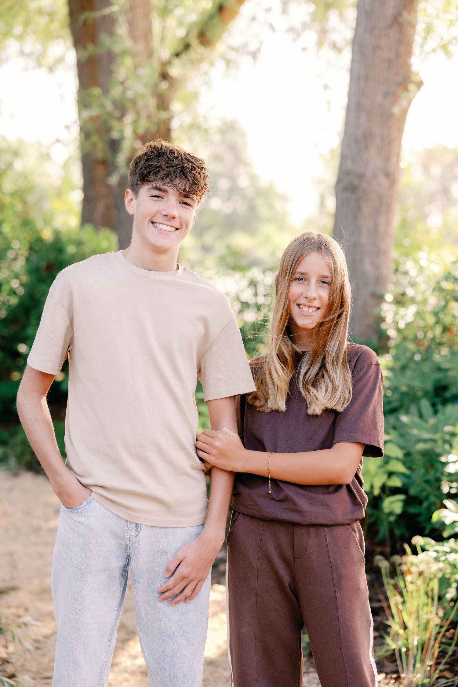 Two smiling teenagers in casual neutral outfits stand side by side outdoors among green trees and shrubs in soft golden sunlight.
