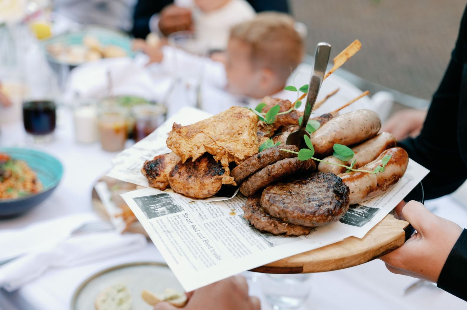 A wooden serving board with grilled meats and sandwiches being served at a restaurant table.