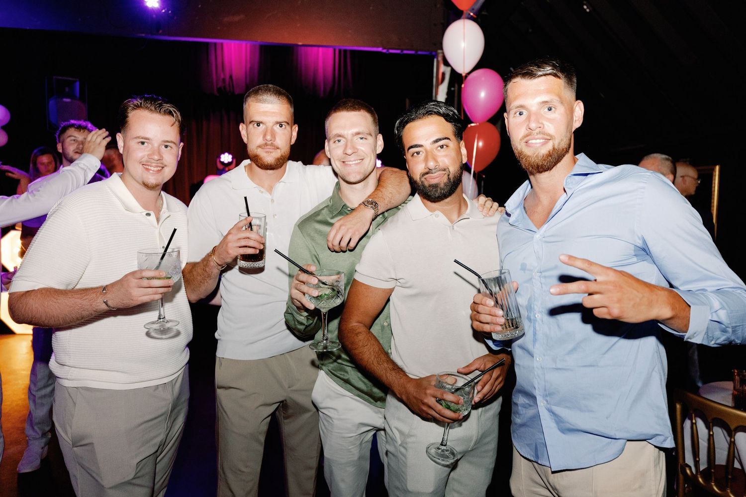 A group of friends enjoying drinks and socializing together at a nightclub with purple lighting and balloons in the background.
