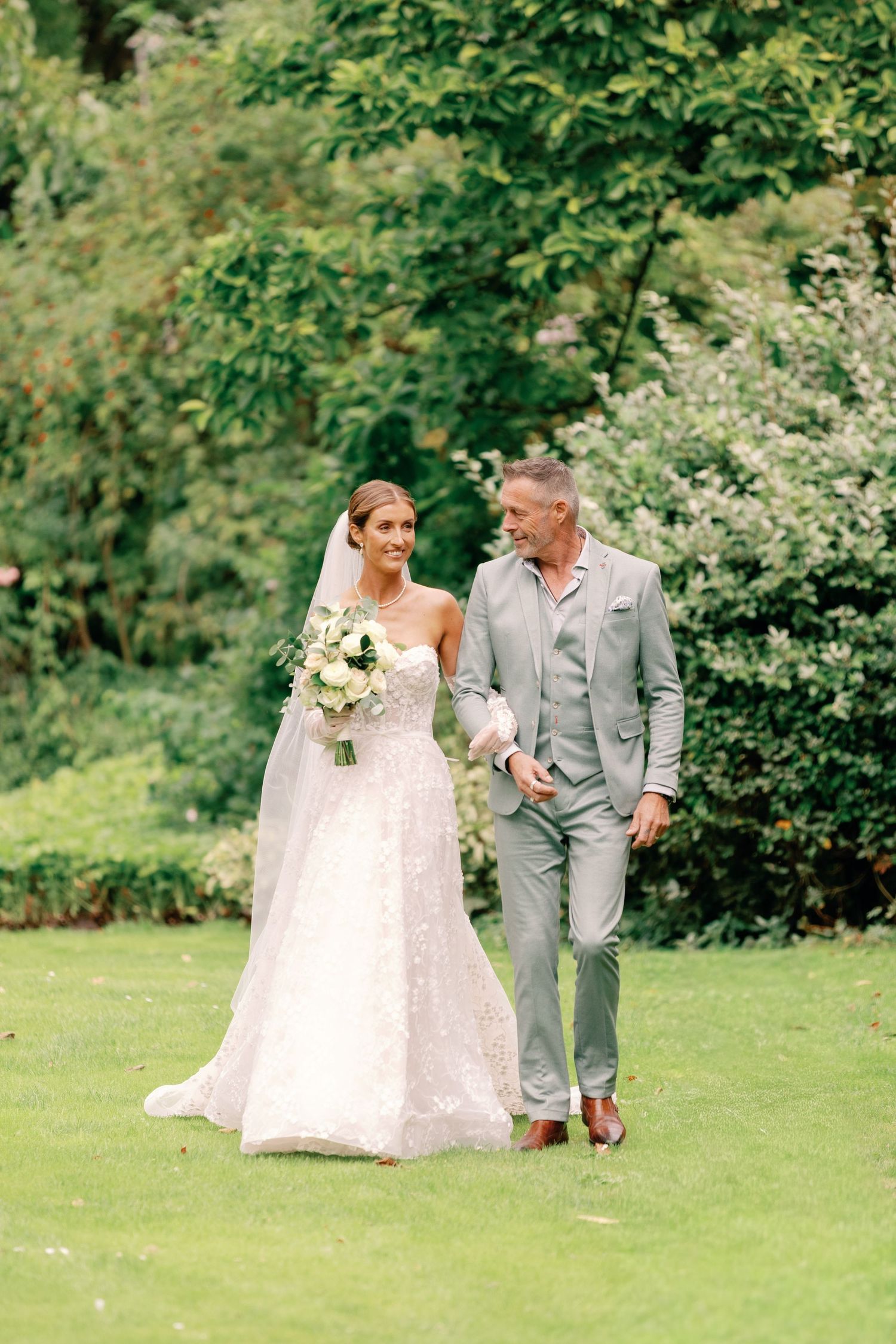 A couple walks together on a lush green lawn during an outdoor wedding ceremony, with the bride in white and groom in gray.