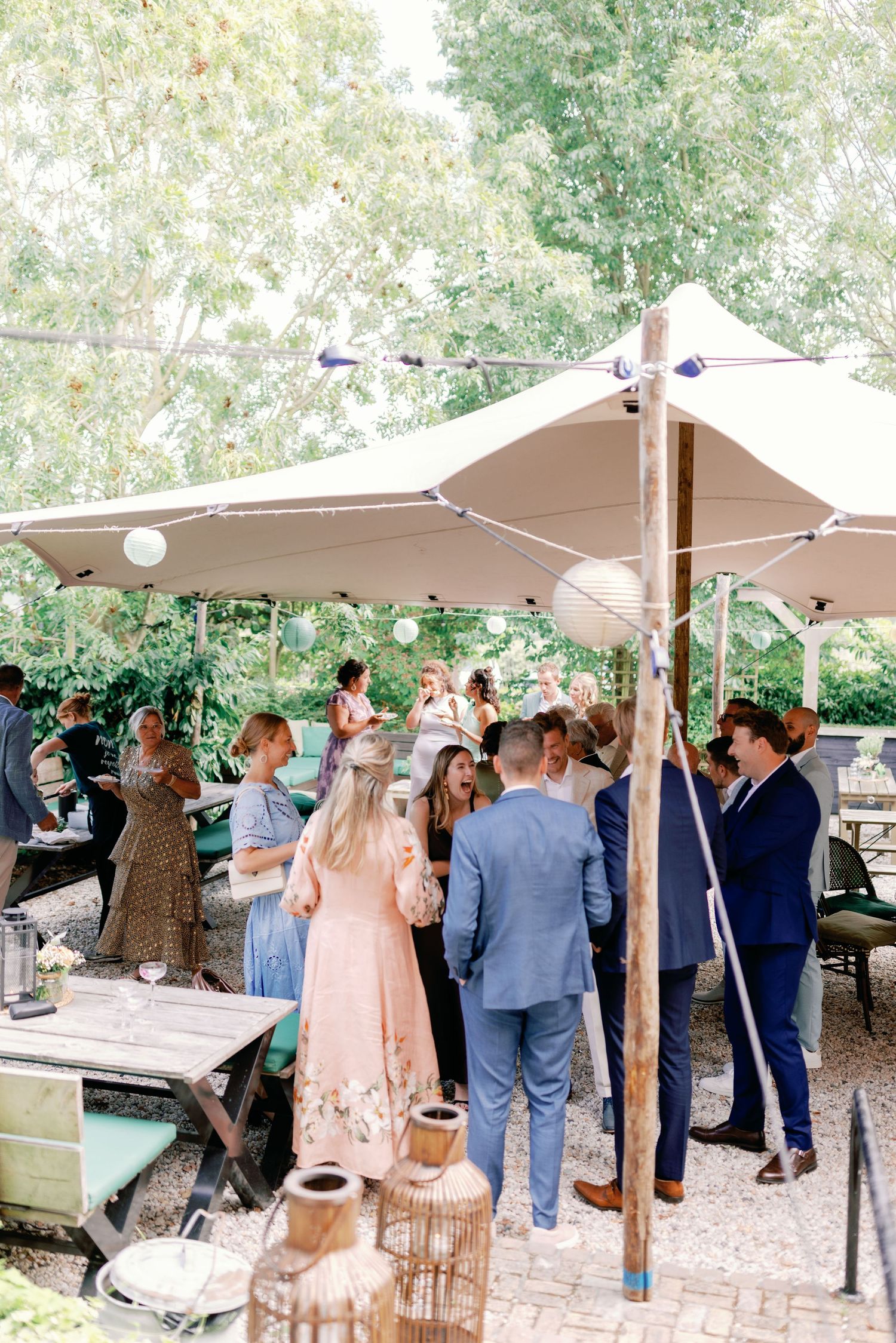 Guests gather under a beige canopy tent at an outdoor event with decorative vases and greenery in the background.