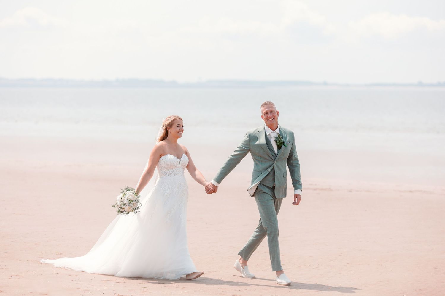 Couple walking hand in hand along a peaceful beach in wedding attire.