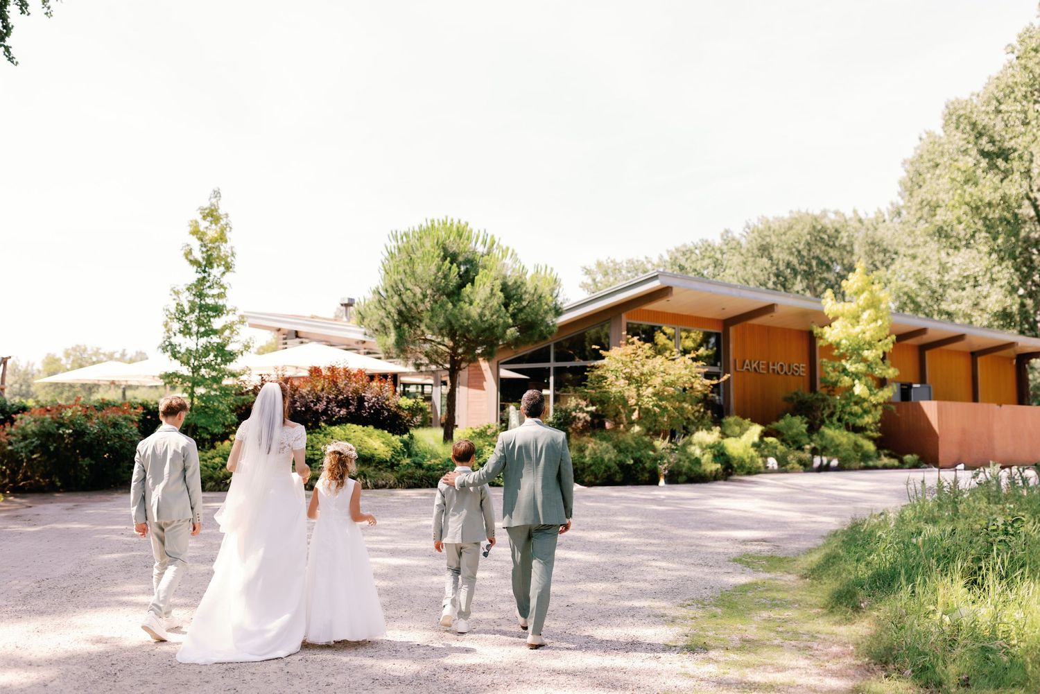 Wedding party walks along garden path outside rustic venue on sunny day.