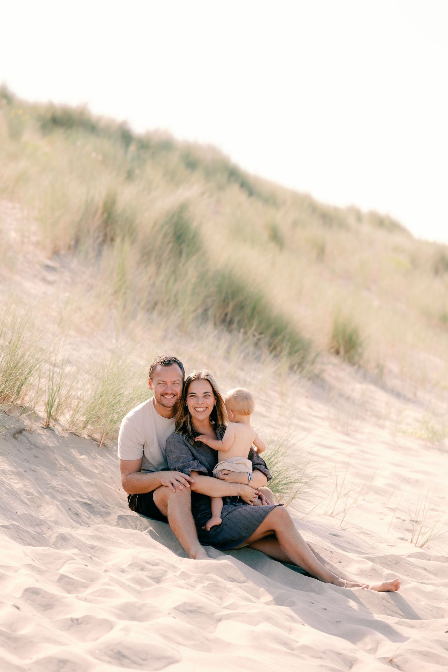 A happy couple sits on sandy beach dunes with their baby, smiling at the camera with grass hills behind them.