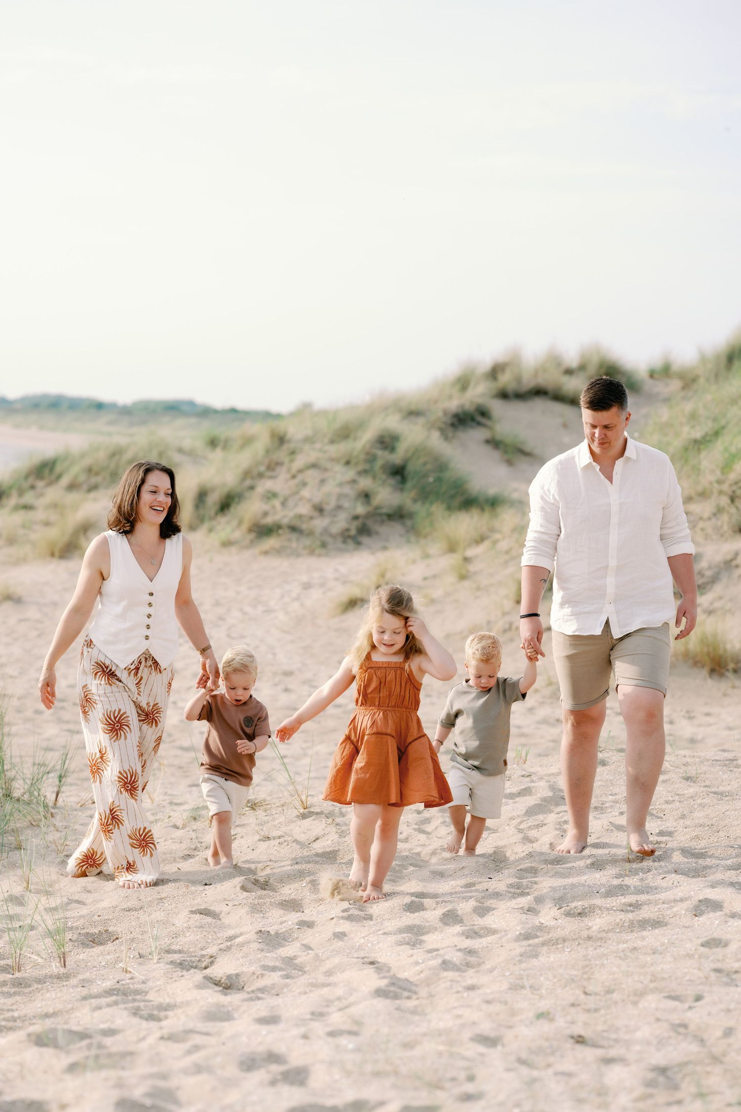 Happy family of five walking hand-in-hand along sandy beach dunes on a bright sunny day.