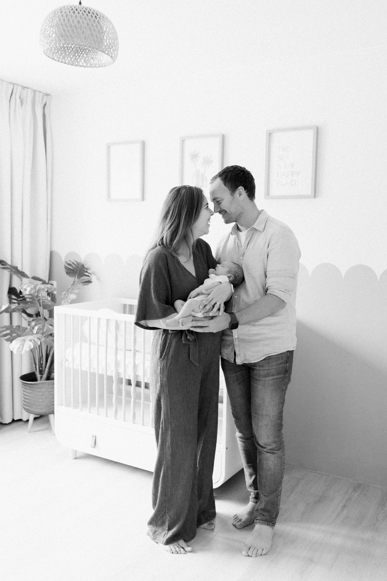 A couple holds their newborn baby in a bright white nursery with a modern pendant light and crib.