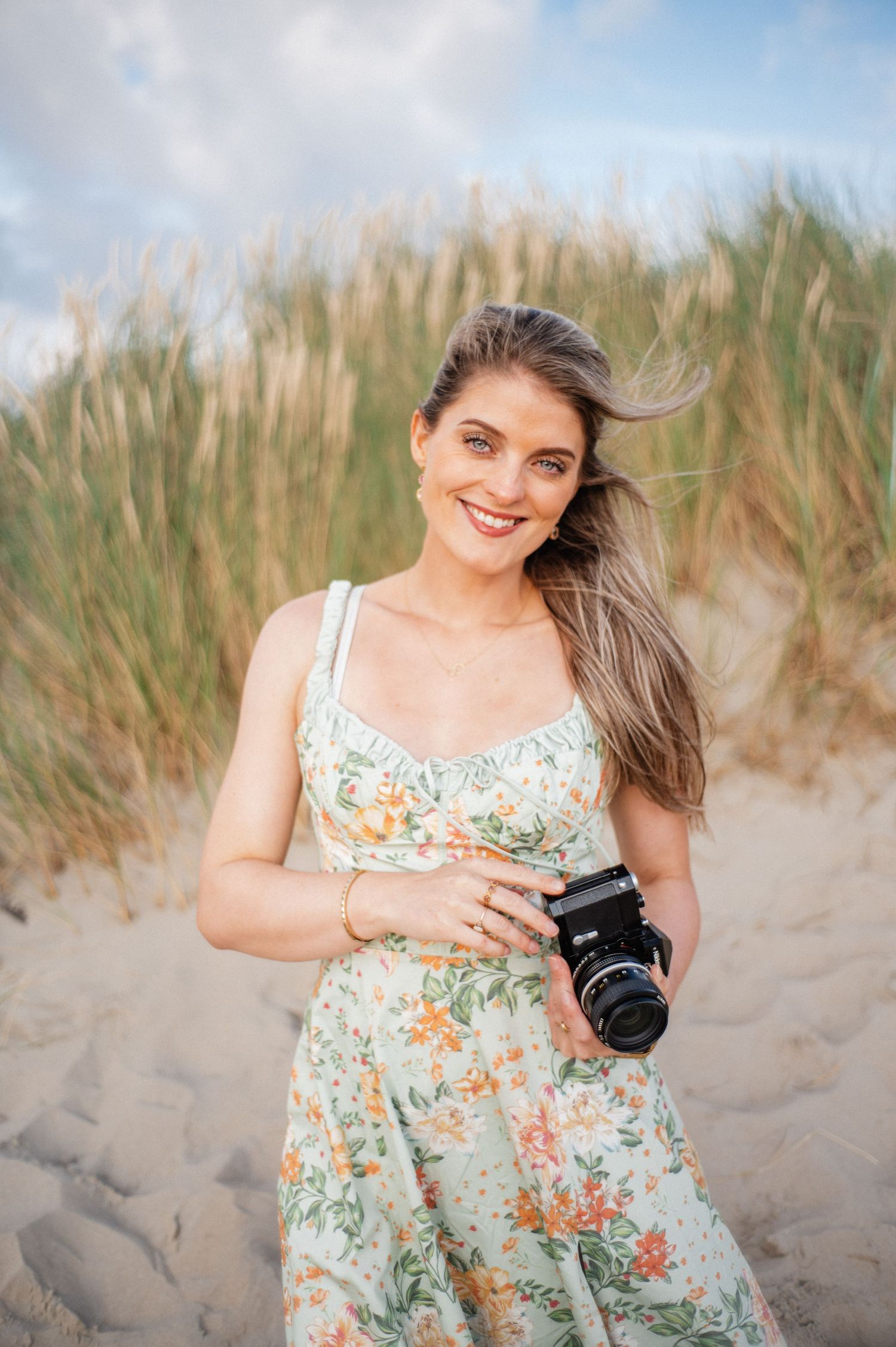 A person in a floral sundress sits on sandy beach dunes with tall grass swaying in the background.