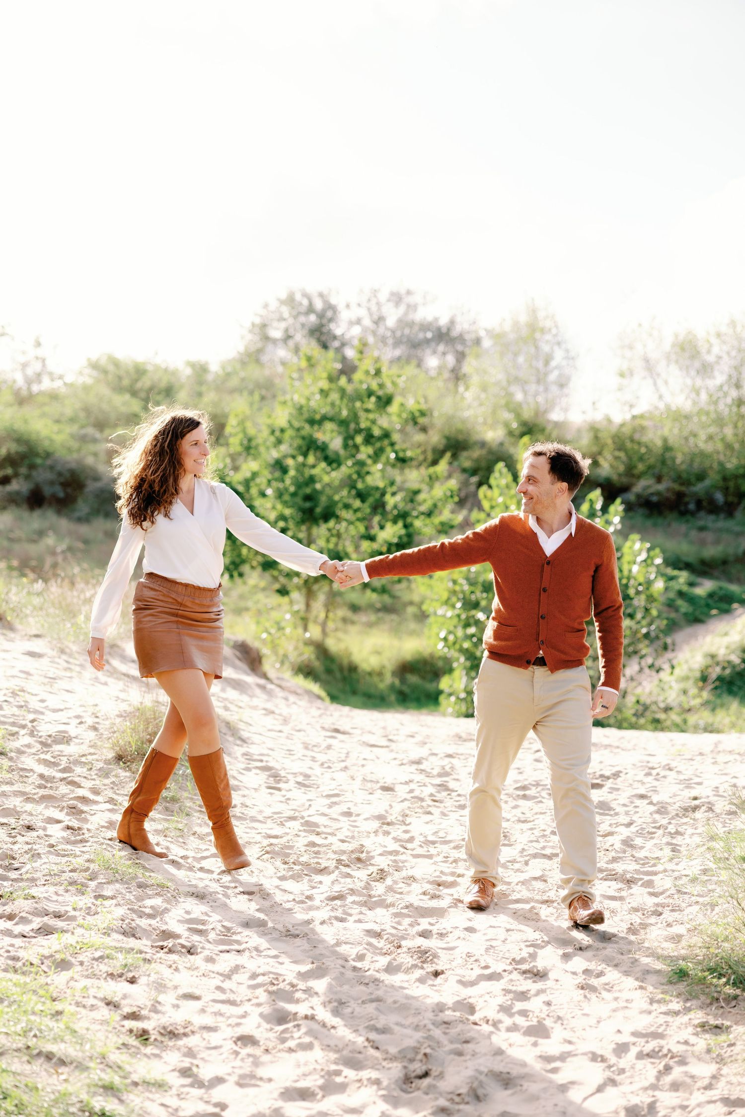 A couple holds hands and walks on a sandy path surrounded by green trees on a sunny day.