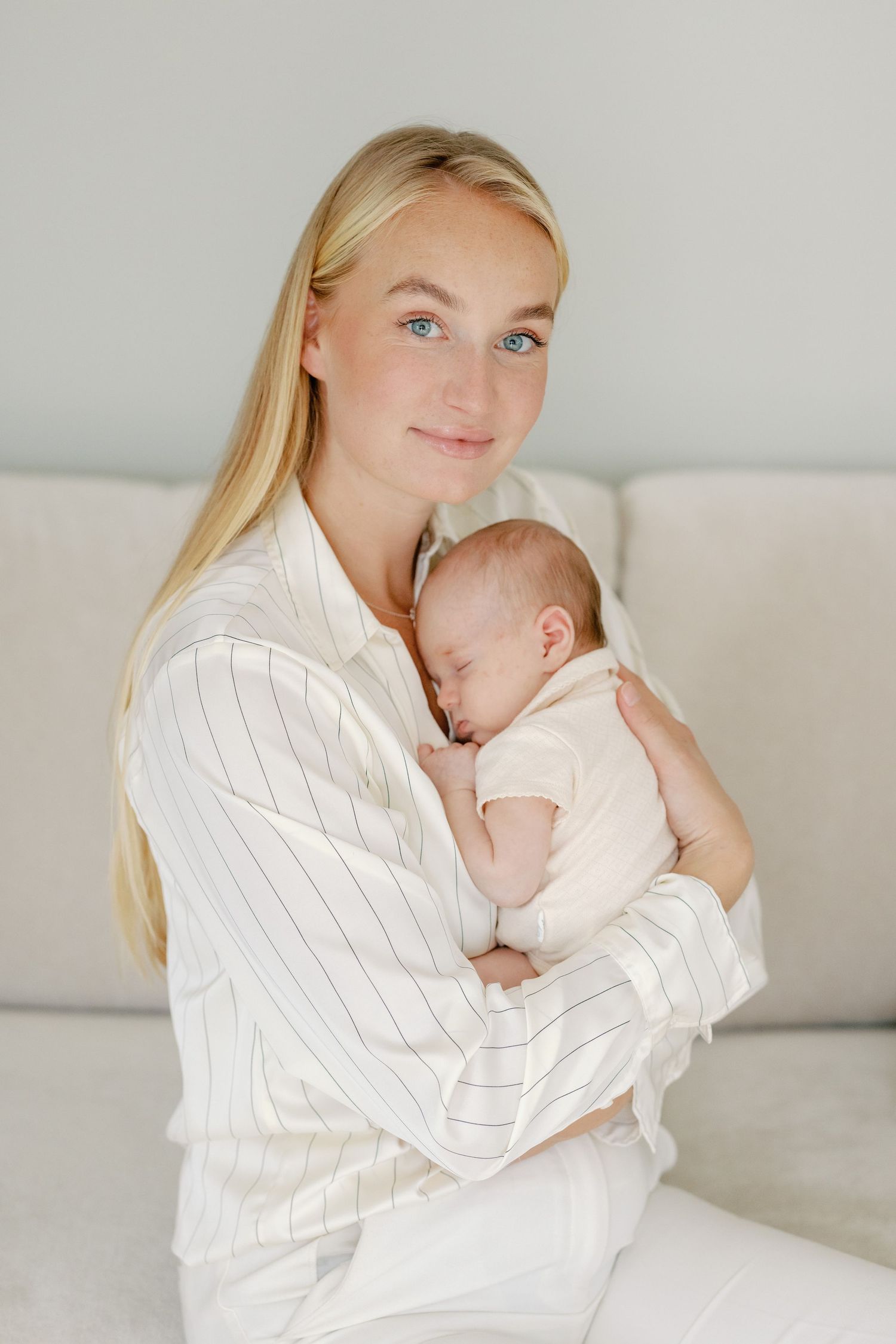 Mother in white pinstripe shirt holds sleeping newborn against neutral background.