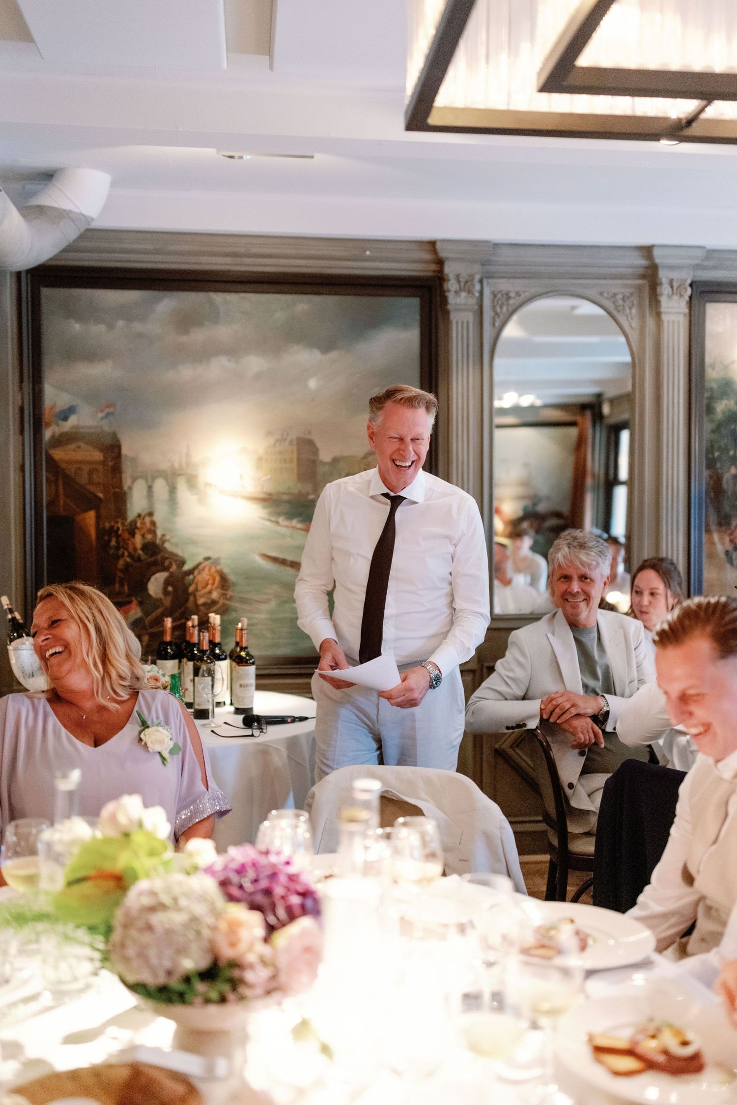 Guests enjoying a formal dinner party at an elegant table decorated with floral centerpieces in a room with landscape paintings.