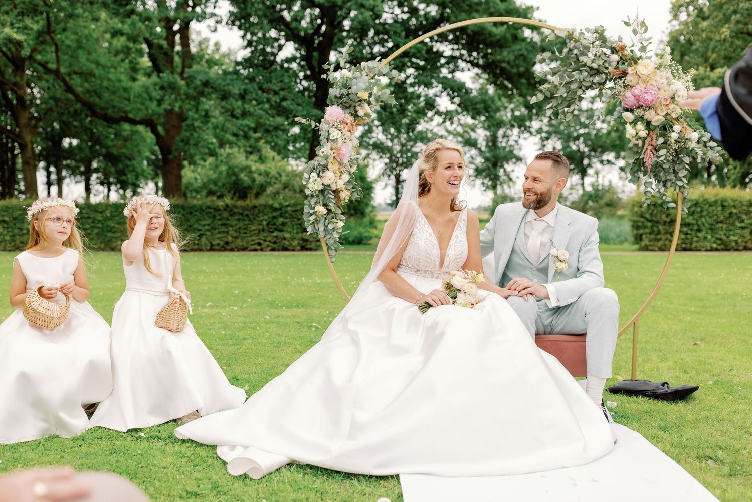 Wedding couple sits on a blanket in a lush green garden setting surrounded by trees and flowers.