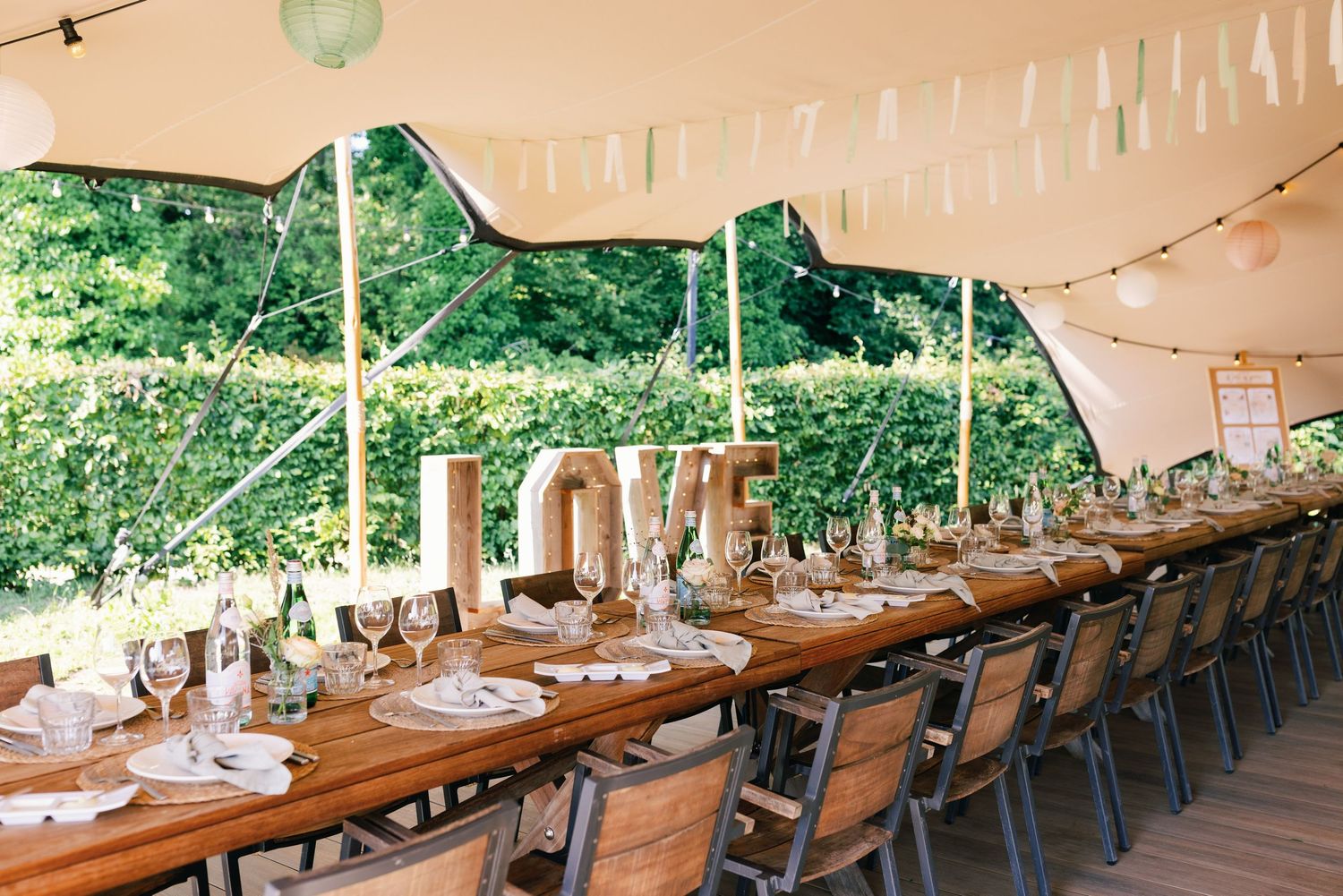 Long wooden tables and chairs set up under a white tent canopy for an elegant outdoor dining event.