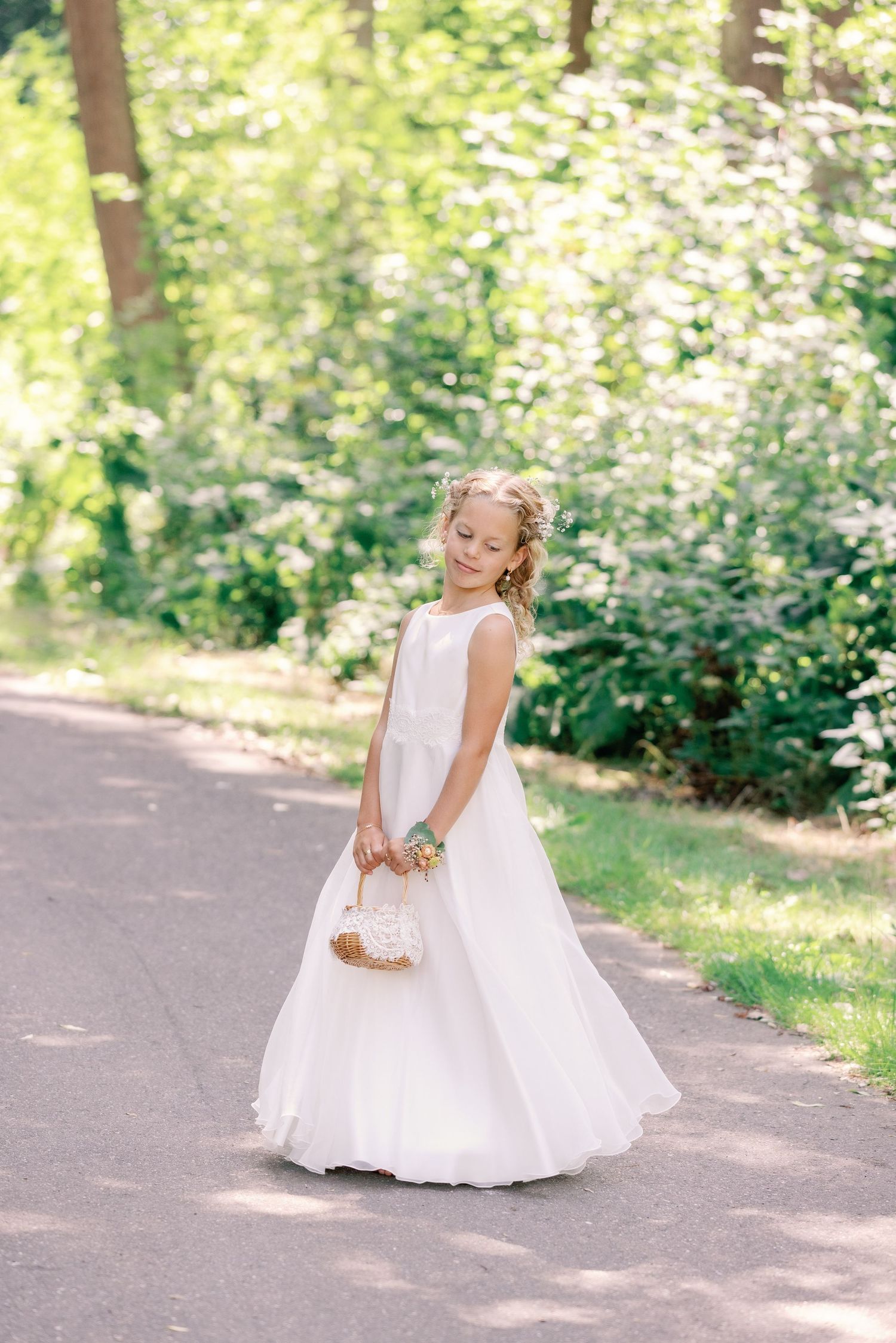 Young flower girl in white dress twirls and dances joyfully on a sunlit garden path during a wedding celebration.