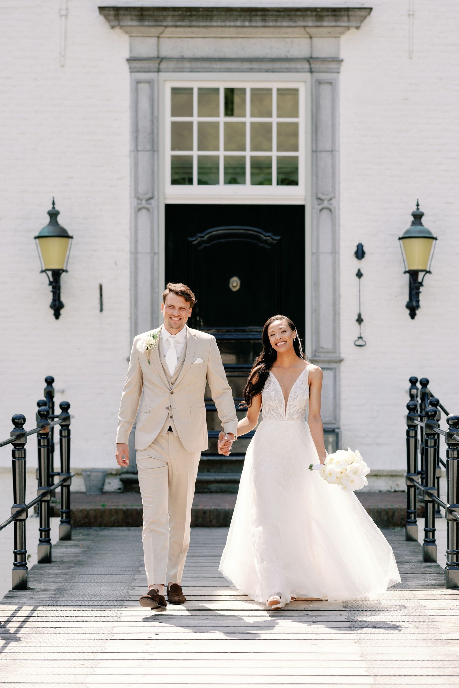 Newlywed couple walks down white building steps with elegant black railings and vintage lanterns on wedding day.