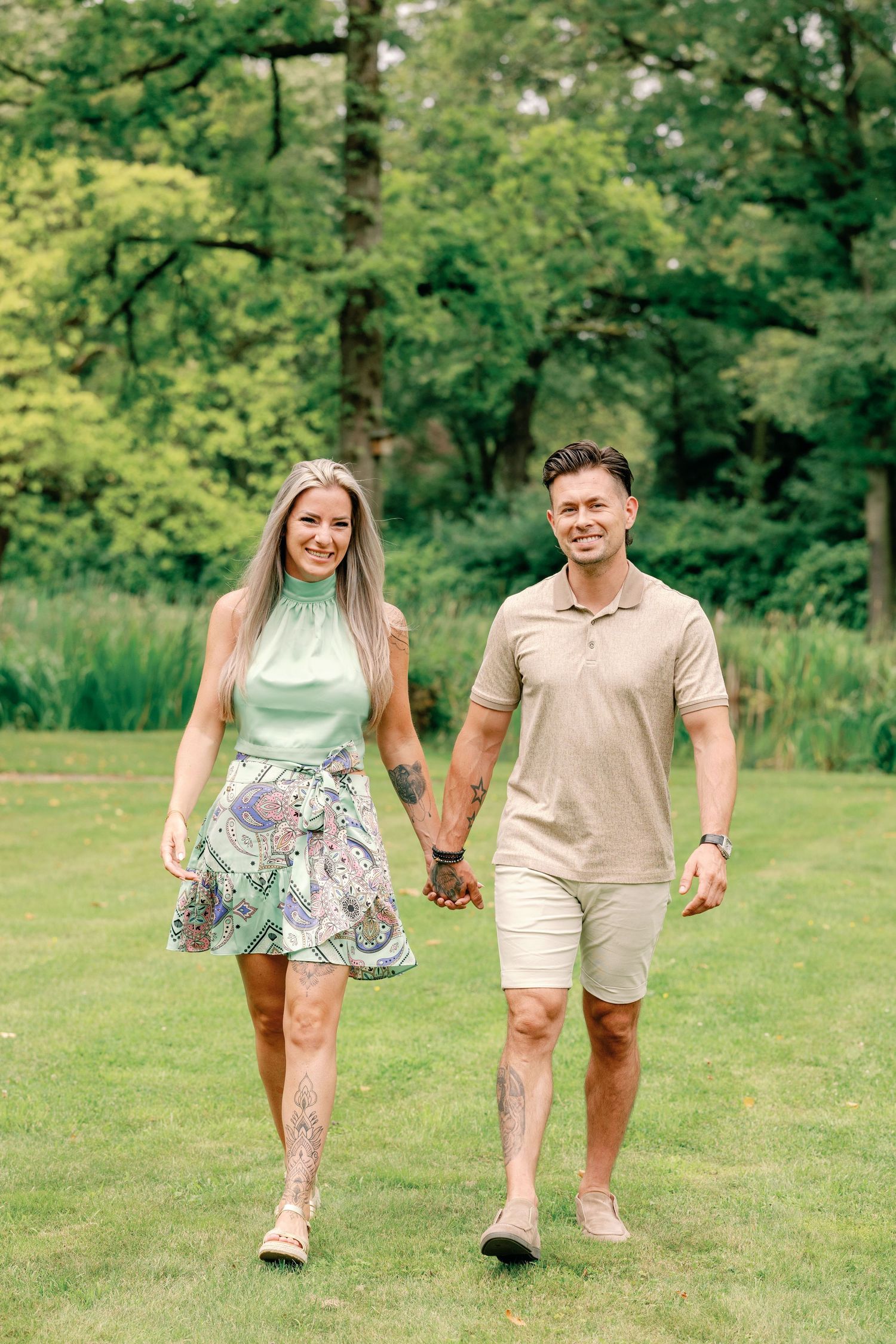 A couple walks hand in hand through a lush green park on a sunny summer day wearing coordinated casual outfits.