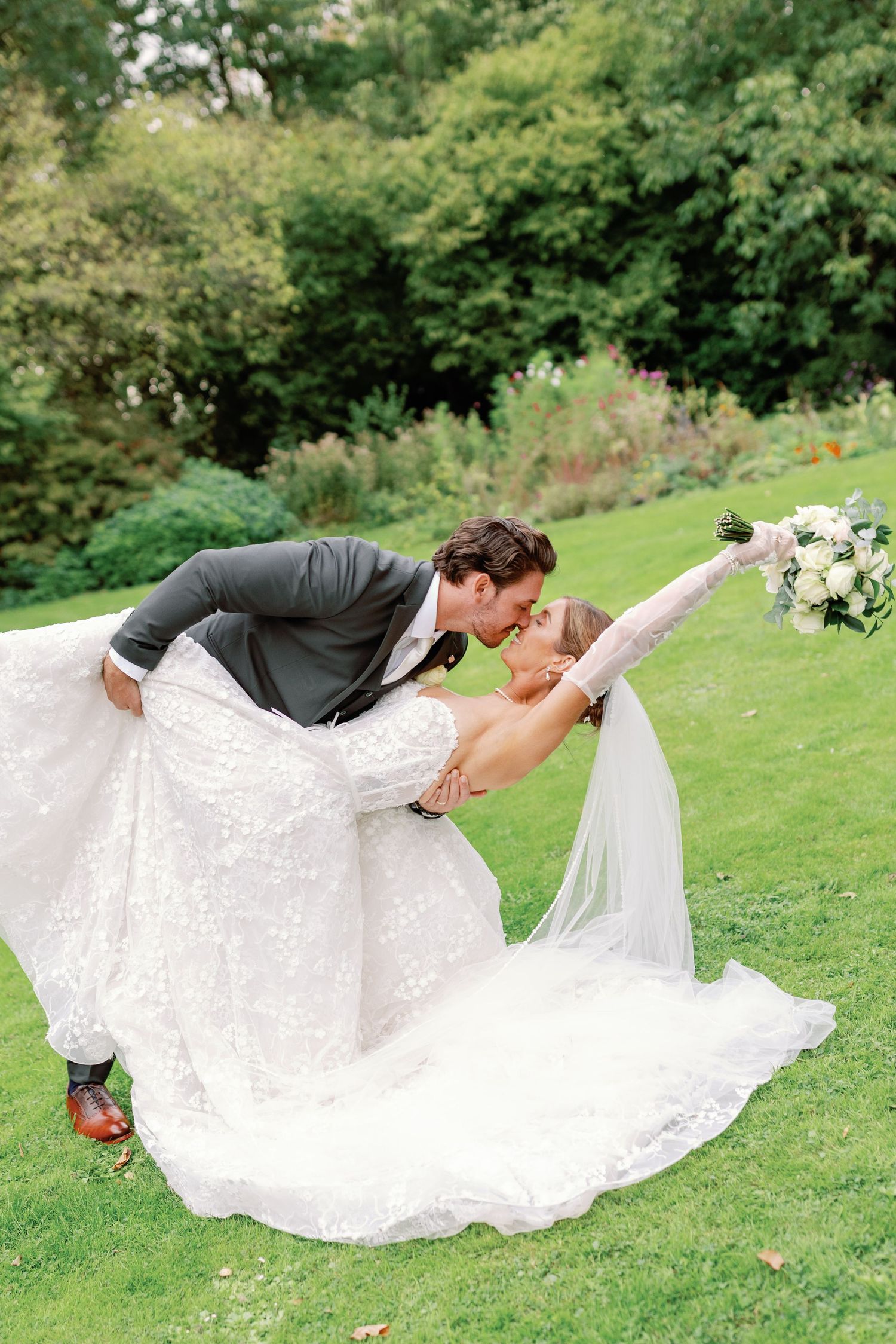 A romantic wedding dip pose on a lush green lawn with the bride in a flowing white lace gown.