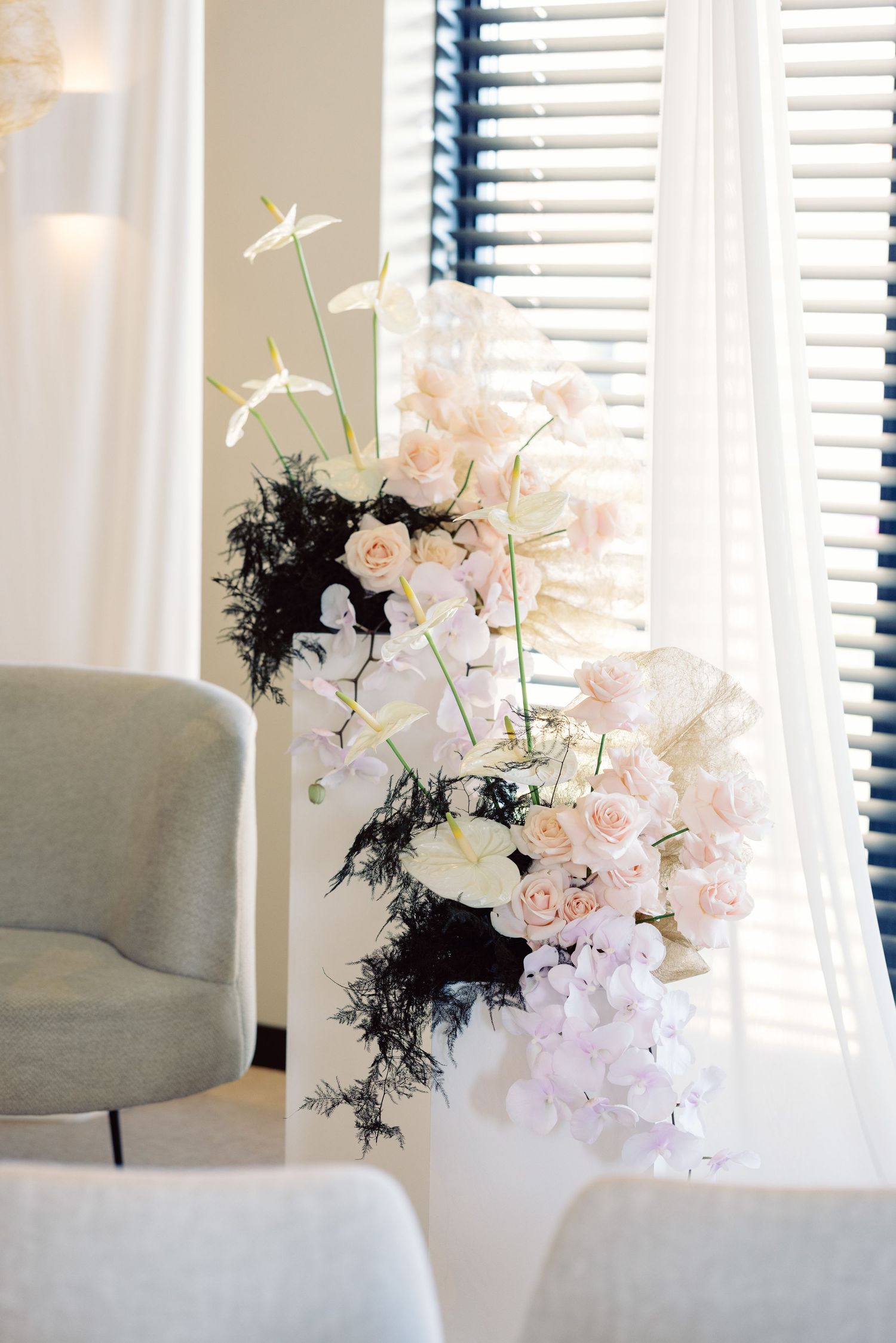Elegant white floral arrangement with orchids near window with venetian blinds.