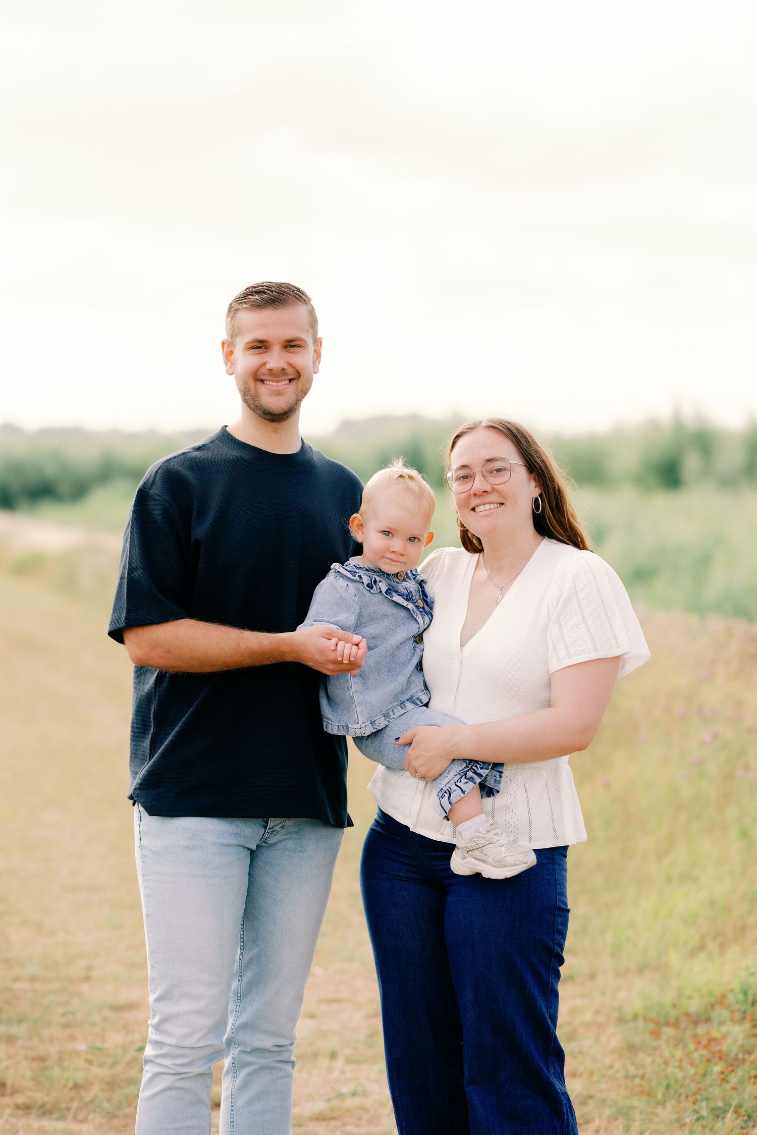 A young family of three poses on a rural path, with parents smiling and holding their baby dressed in denim.