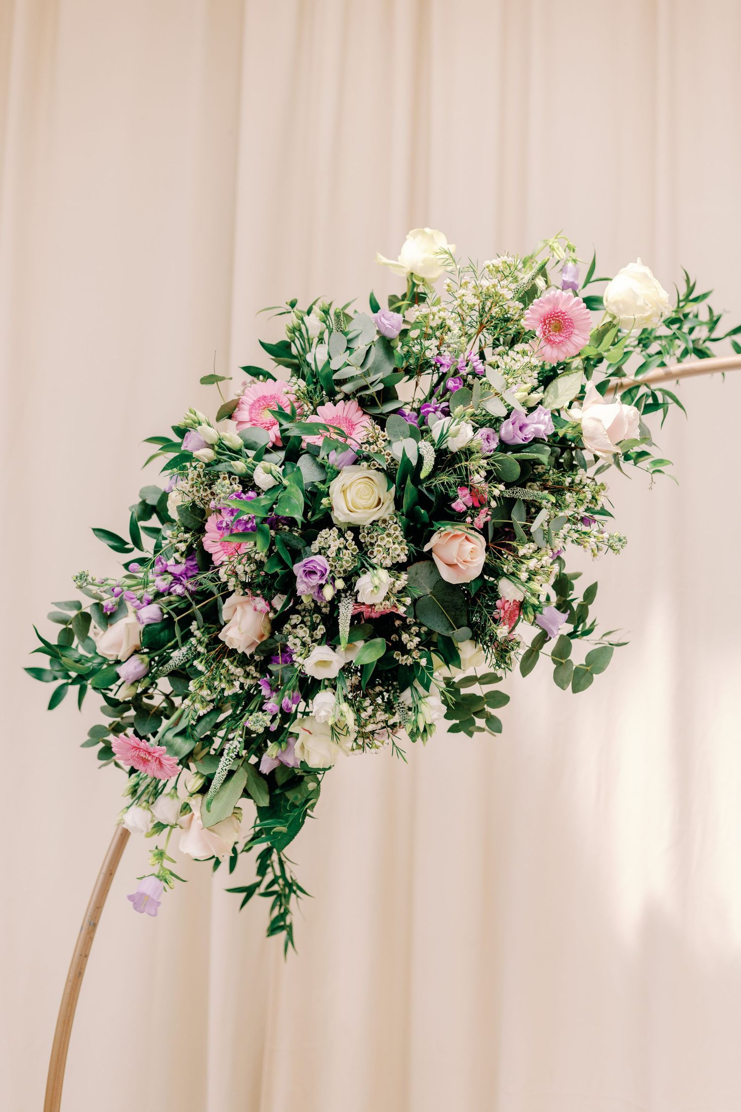 Romantic floral arrangement with pink and purple blooms mixed with greenery hanging against a light fabric backdrop.