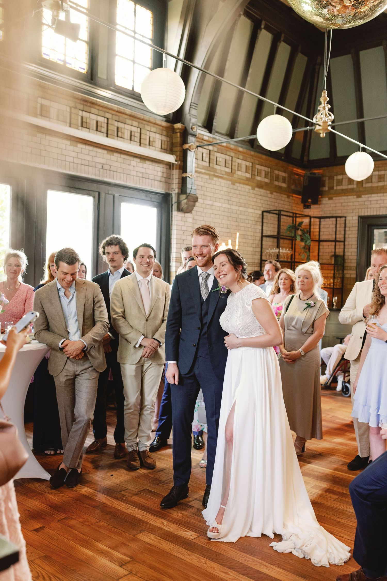 Wedding guests line up along the wall of an elegant brick-walled reception venue with hardwood floors and globe lights.
