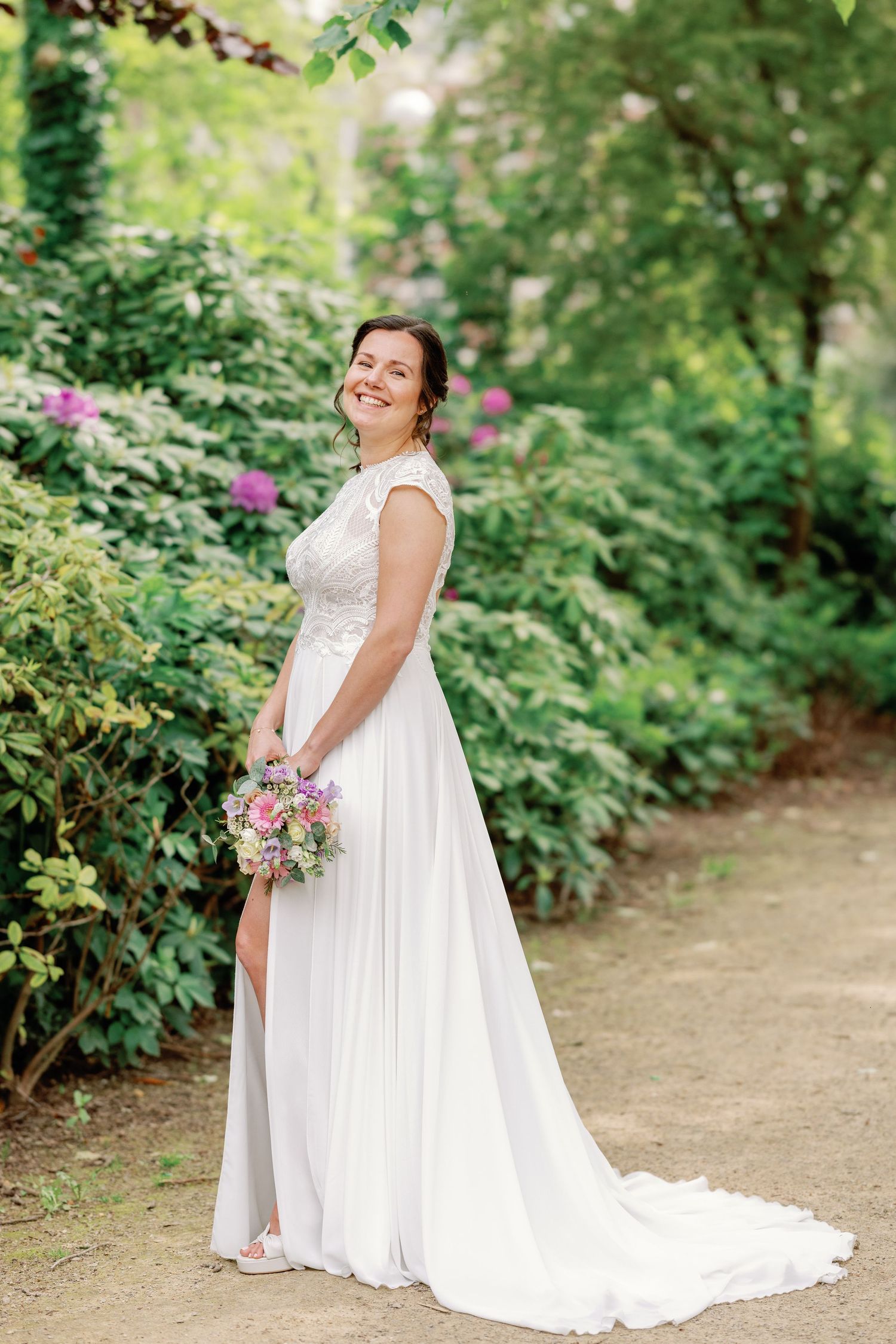 A bride in a flowing white wedding gown poses elegantly in a garden setting with lush greenery and blooming flowers.