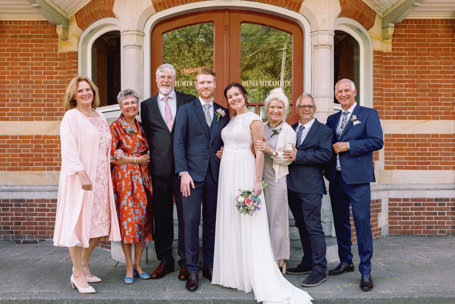 Wedding party members in formal attire stand together outside a brick building with arched windows.