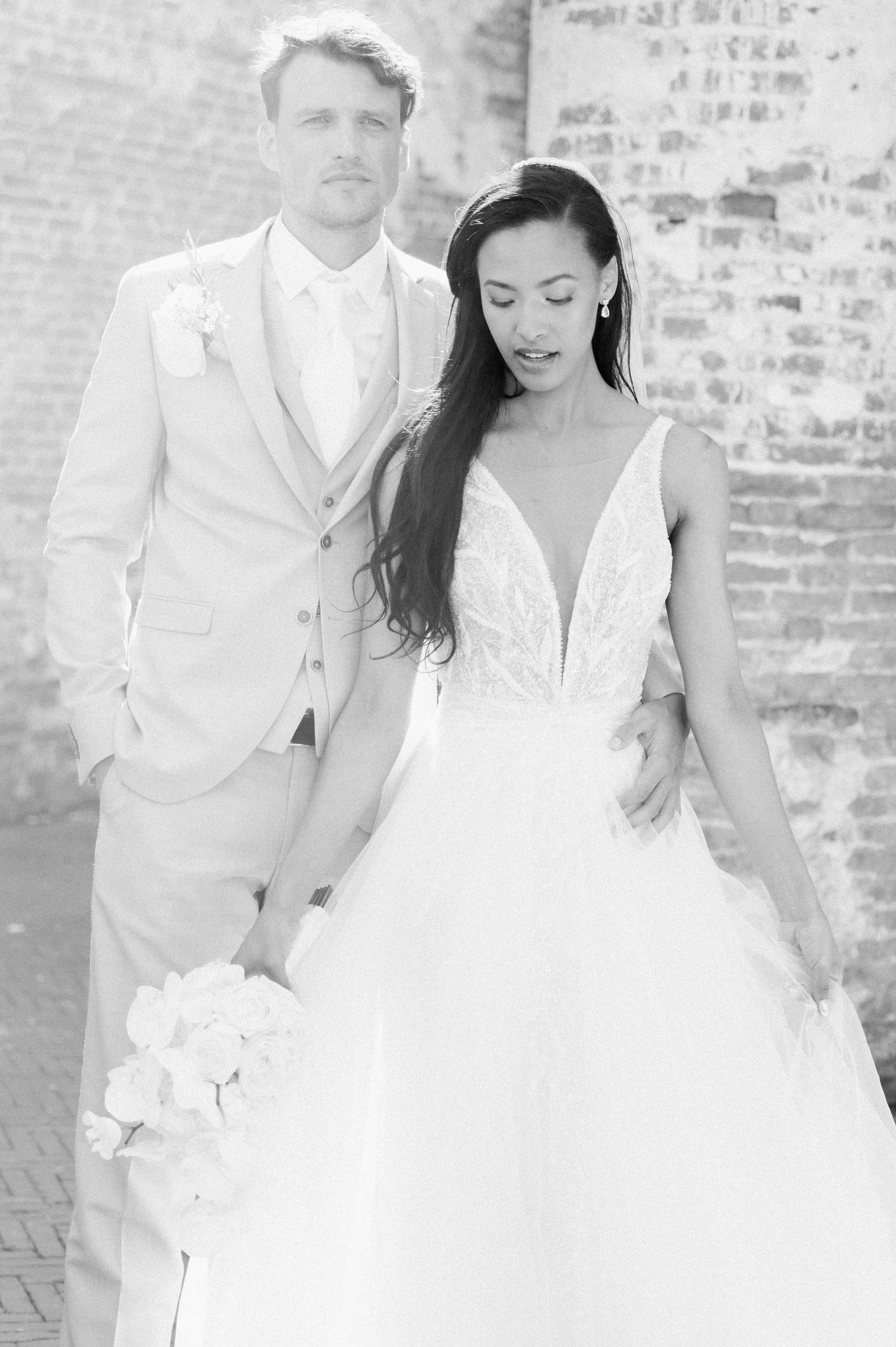 A bride and groom share intimate black and white wedding portraits against a rustic brick wall backdrop.