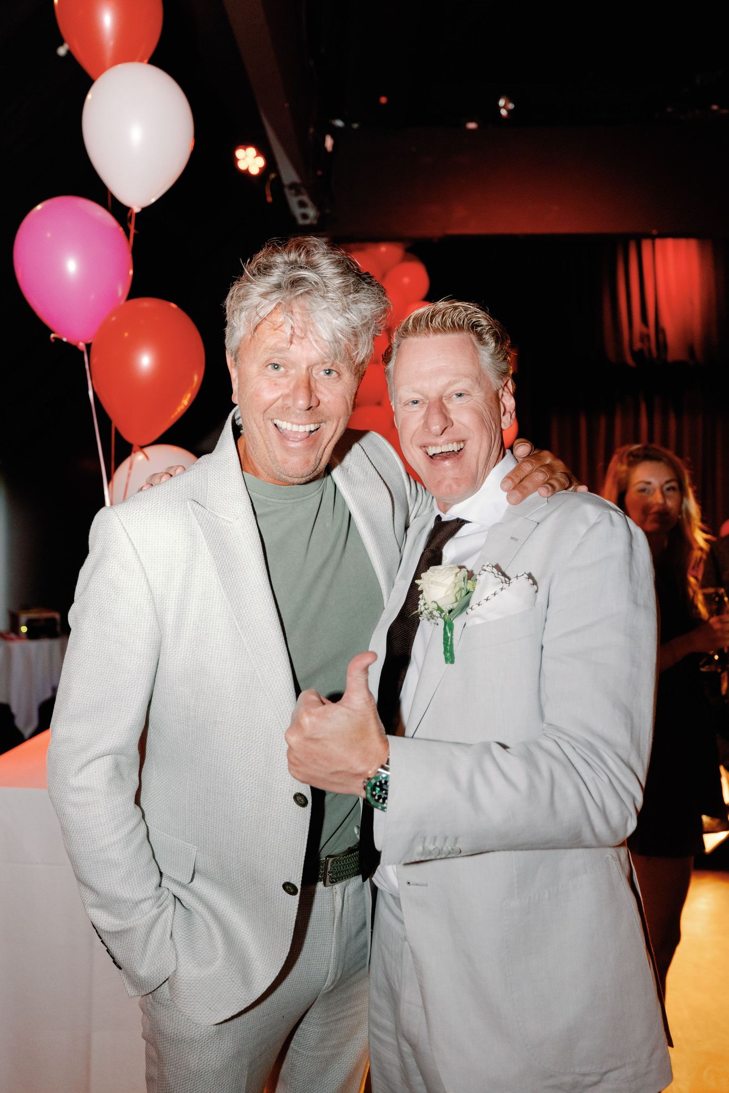 Two people in light colored suits smile and pose together with red and white balloons at an indoor event.