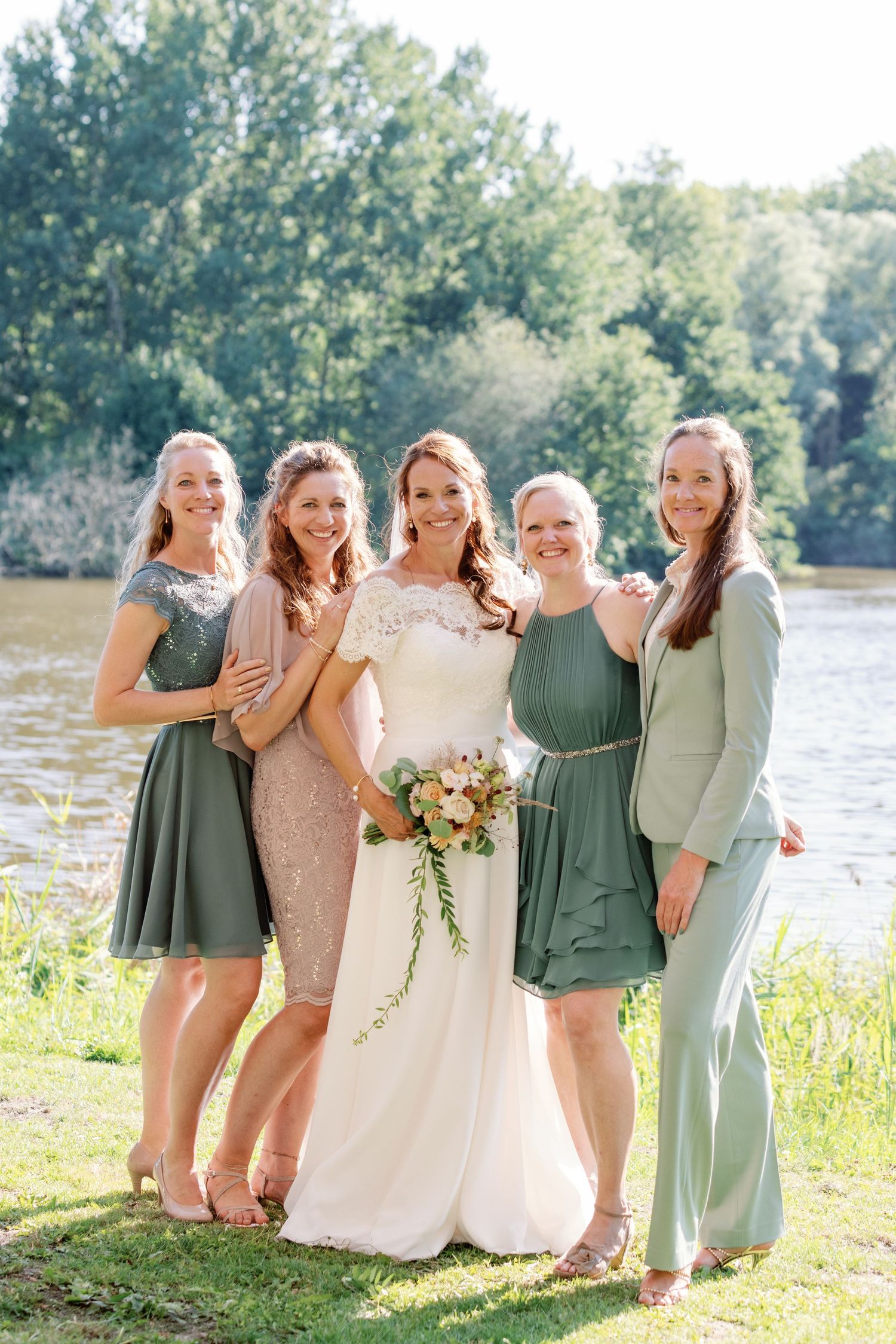 Bridesmaids in sage green dresses laugh together with bride in white gown by lakeside on sunny summer day.